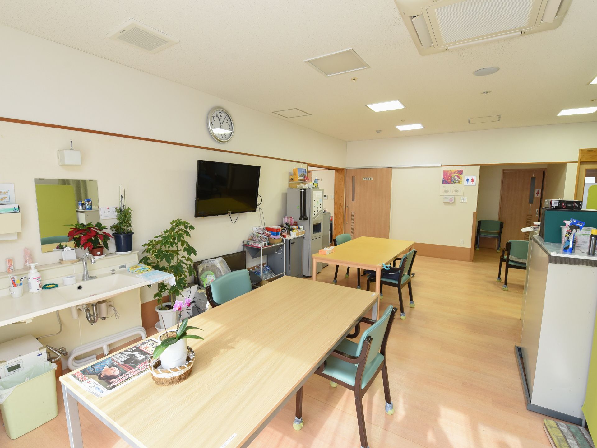 壁掛けテレビの左側に洗面台が設置されている、日差しが差し込む食堂