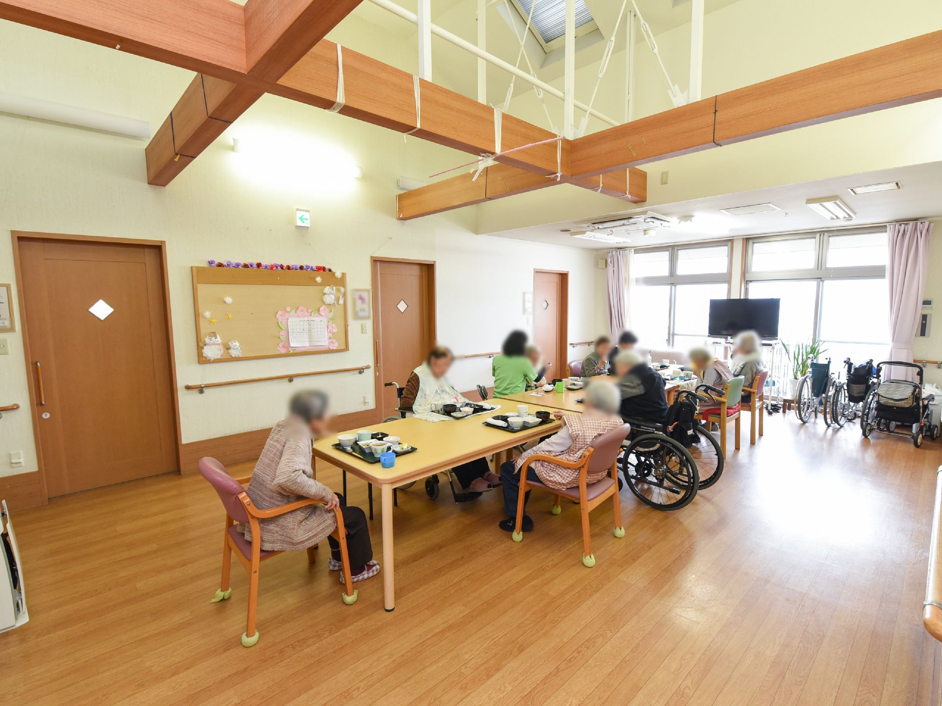 入居者様が食事をしている様子である。大きな窓かあり、明るい光が入ってきている。壁には掲示物や飾りが飾られている。