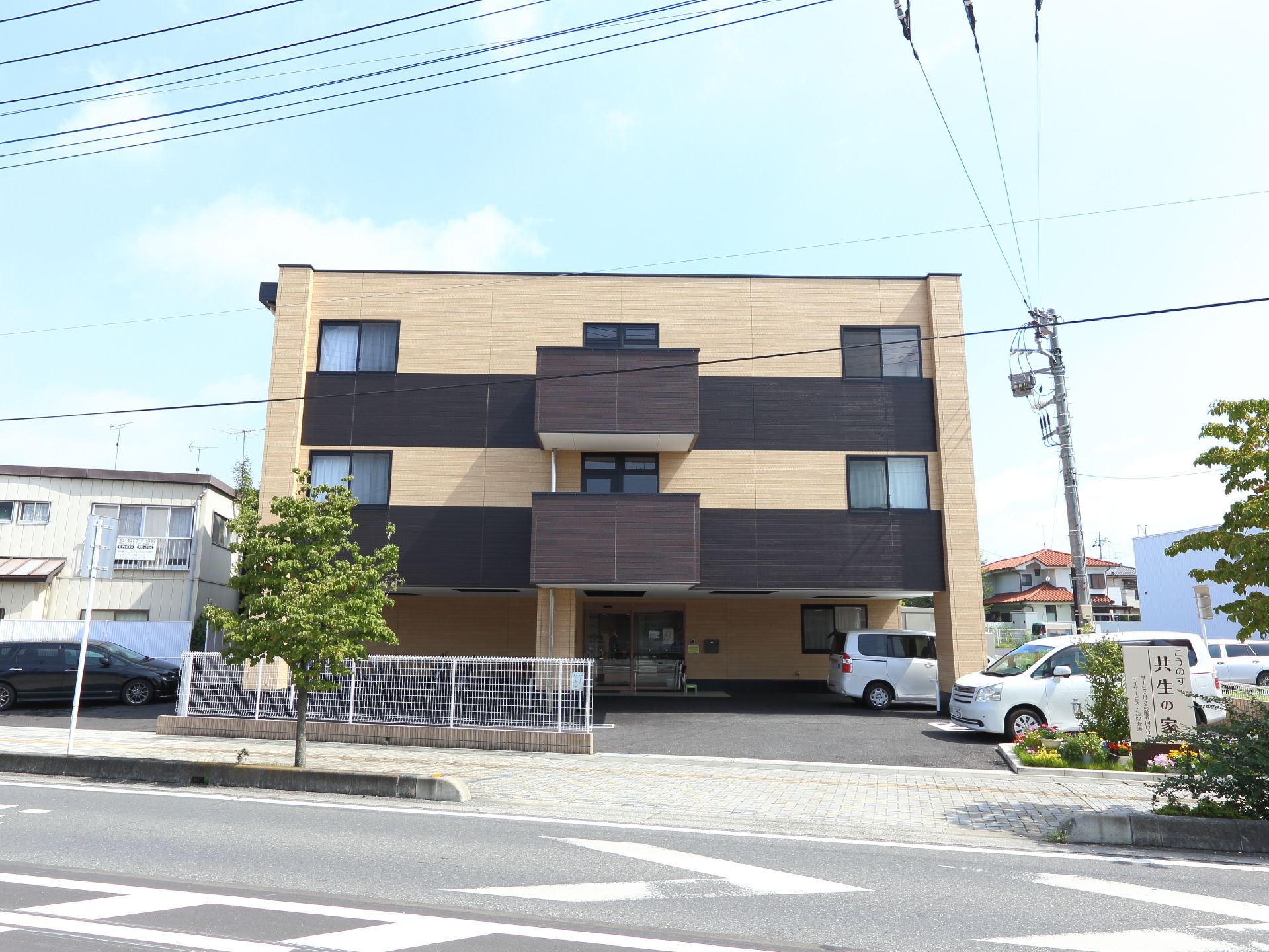 施設の建物を道路越しの正面から見た画像。全部で3階建ての構造なのが見て取れる。右側に建物名の看板が設置されている。