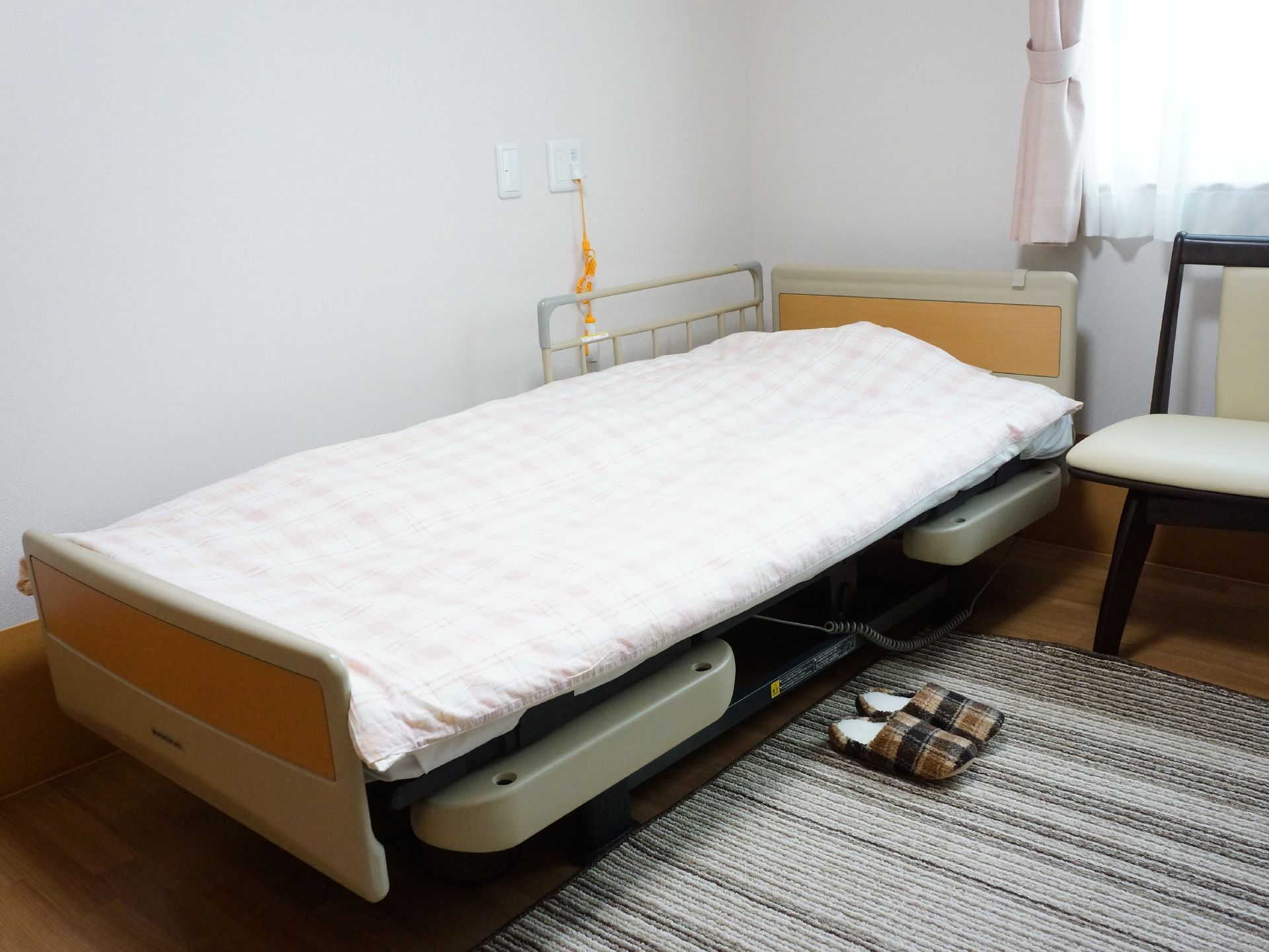 当施設のベッドは電動式である。ベッドには左右に柵がつけられるため、落下防止につながる。奥の壁にはコードのような紐が垂れ下がっている。