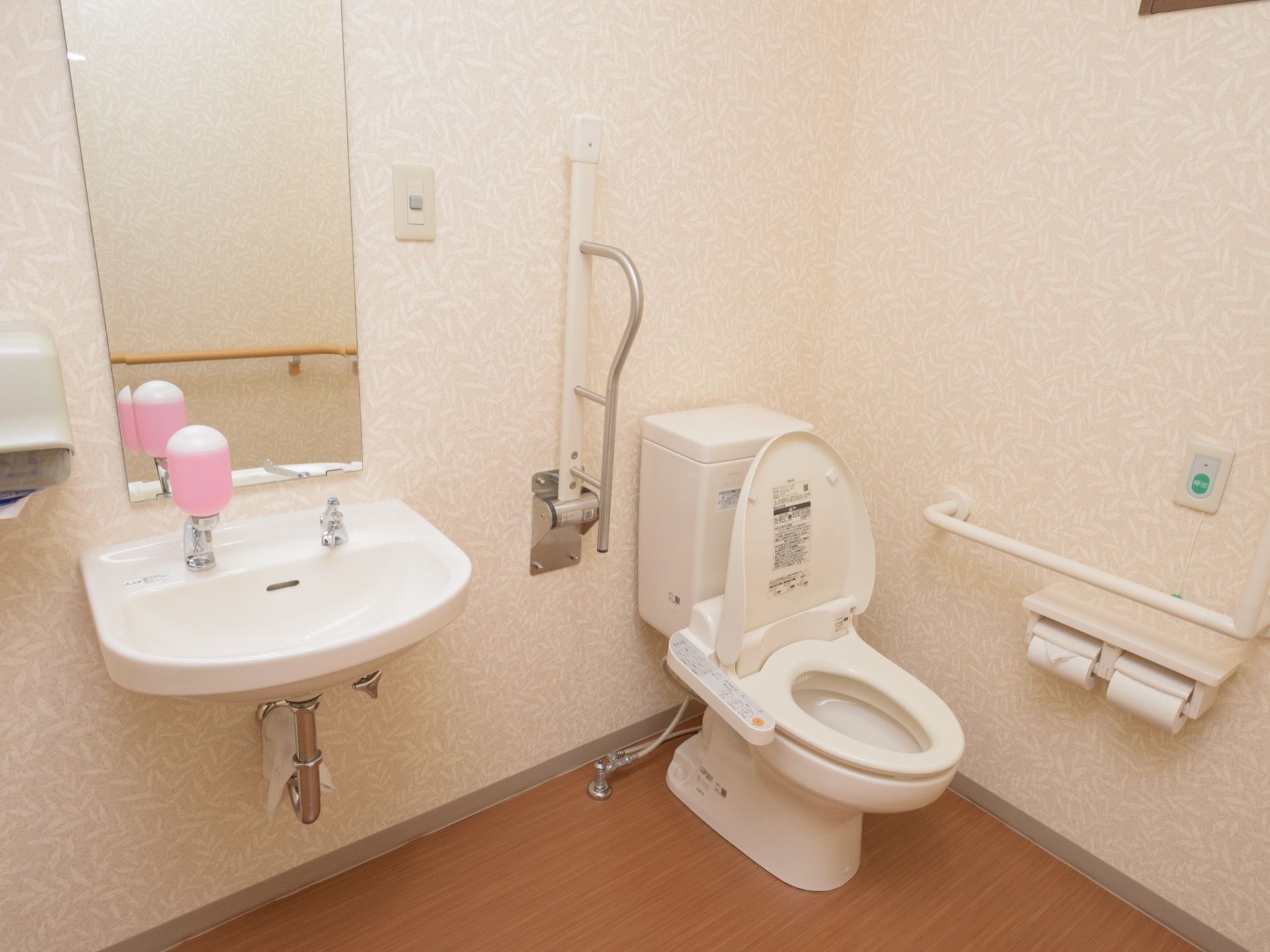 温水洗浄便座付きのトイレである。壁に横長の手すり、反対側に可動式の手すりがある。同じ個室内に、手洗い場も完備されている。