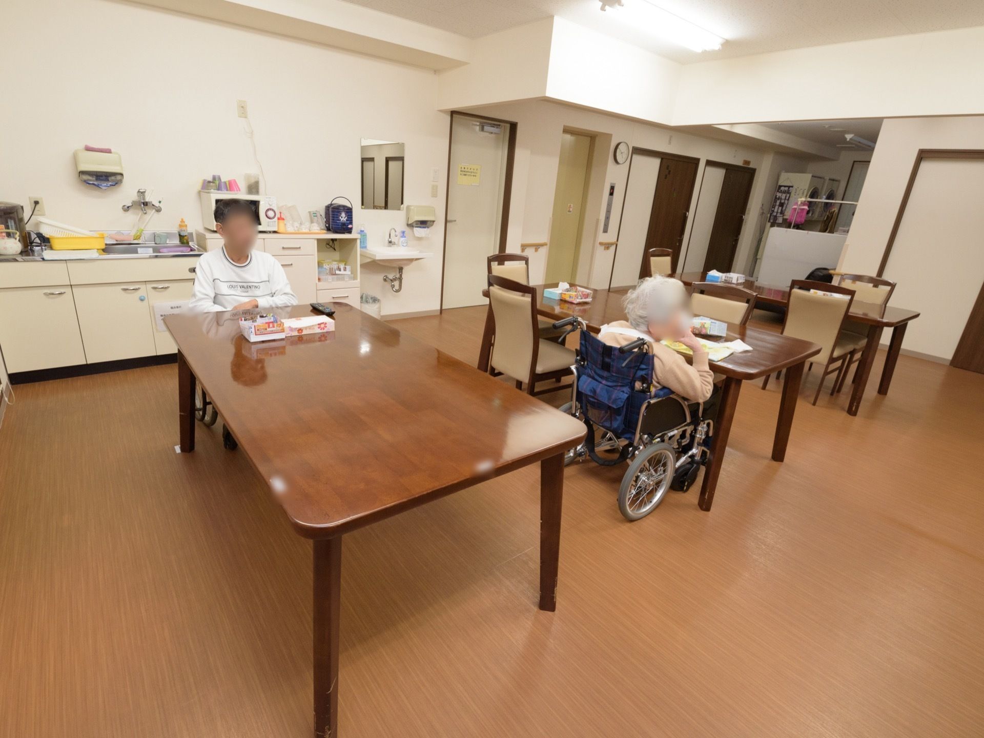 室内に茶色いテーブルセットが3つあり、2人の入居者が席についている。室内の一角にキッチンがあり、電子レンジなどが置かれている。