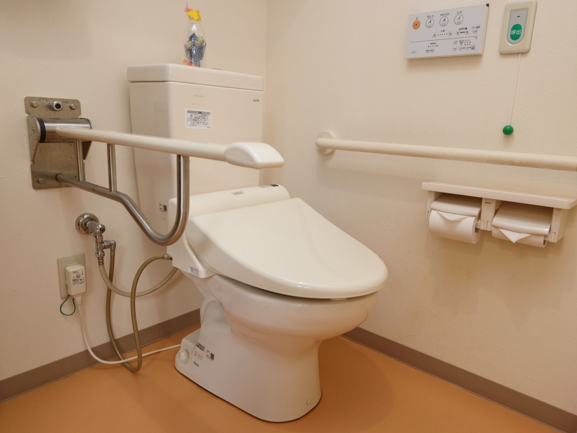 洋式トイレの横に可動式のひじ掛けを設置している。壁にはトイレの操作パネルや手すり、ナースコールが付いている。