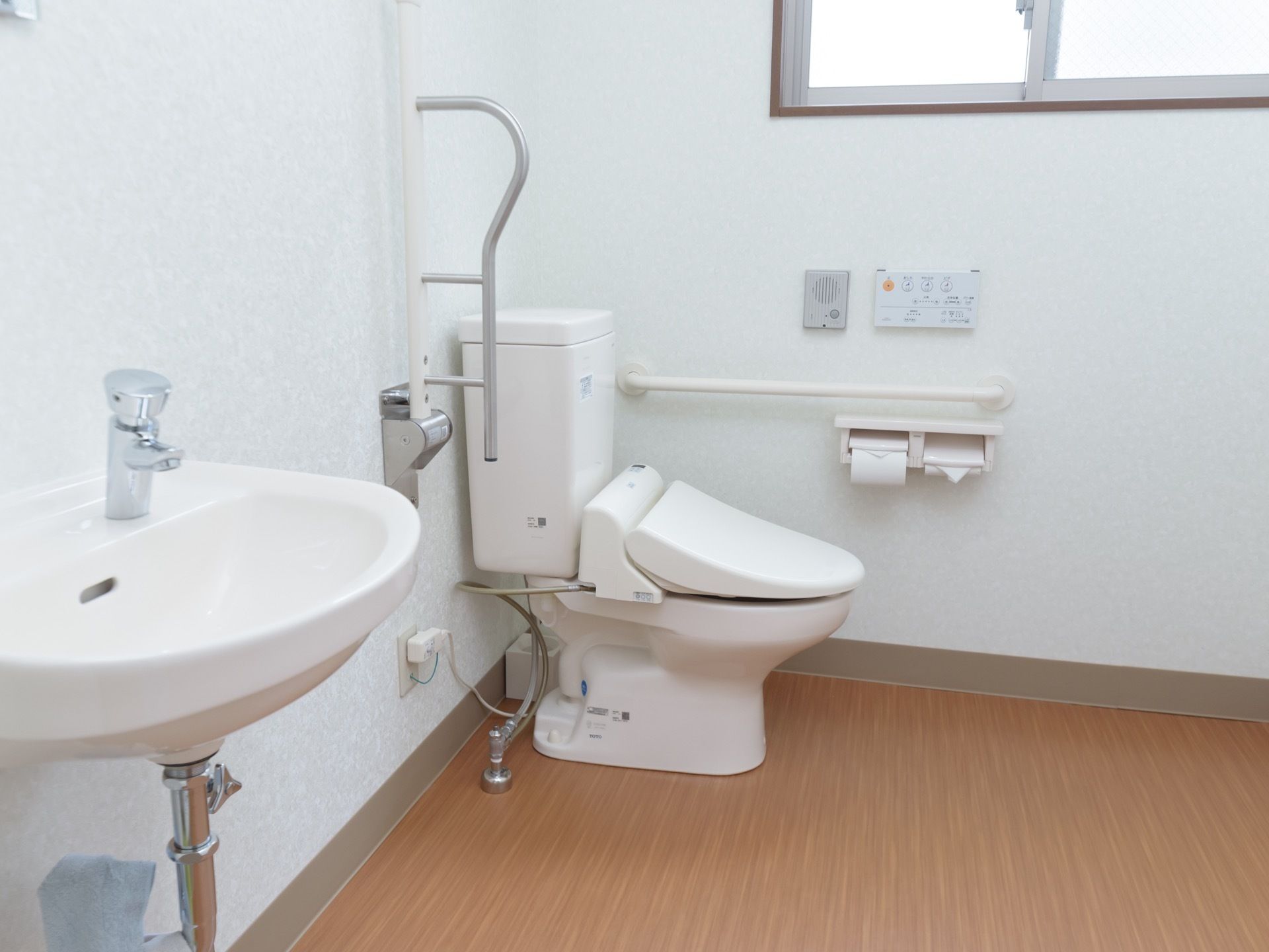 広いトイレである。便座そばの壁に手すりやインターフォンが備わっている。便器、可動式の手すり、洗面台は並んで設置されている。