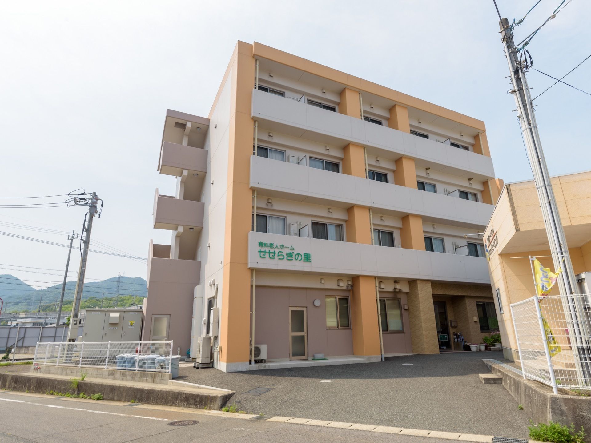 ベージュを基調とした可愛らしい雰囲気の４階建ての建物である。玄関の前に植木鉢の花がいくつか置かれている。
