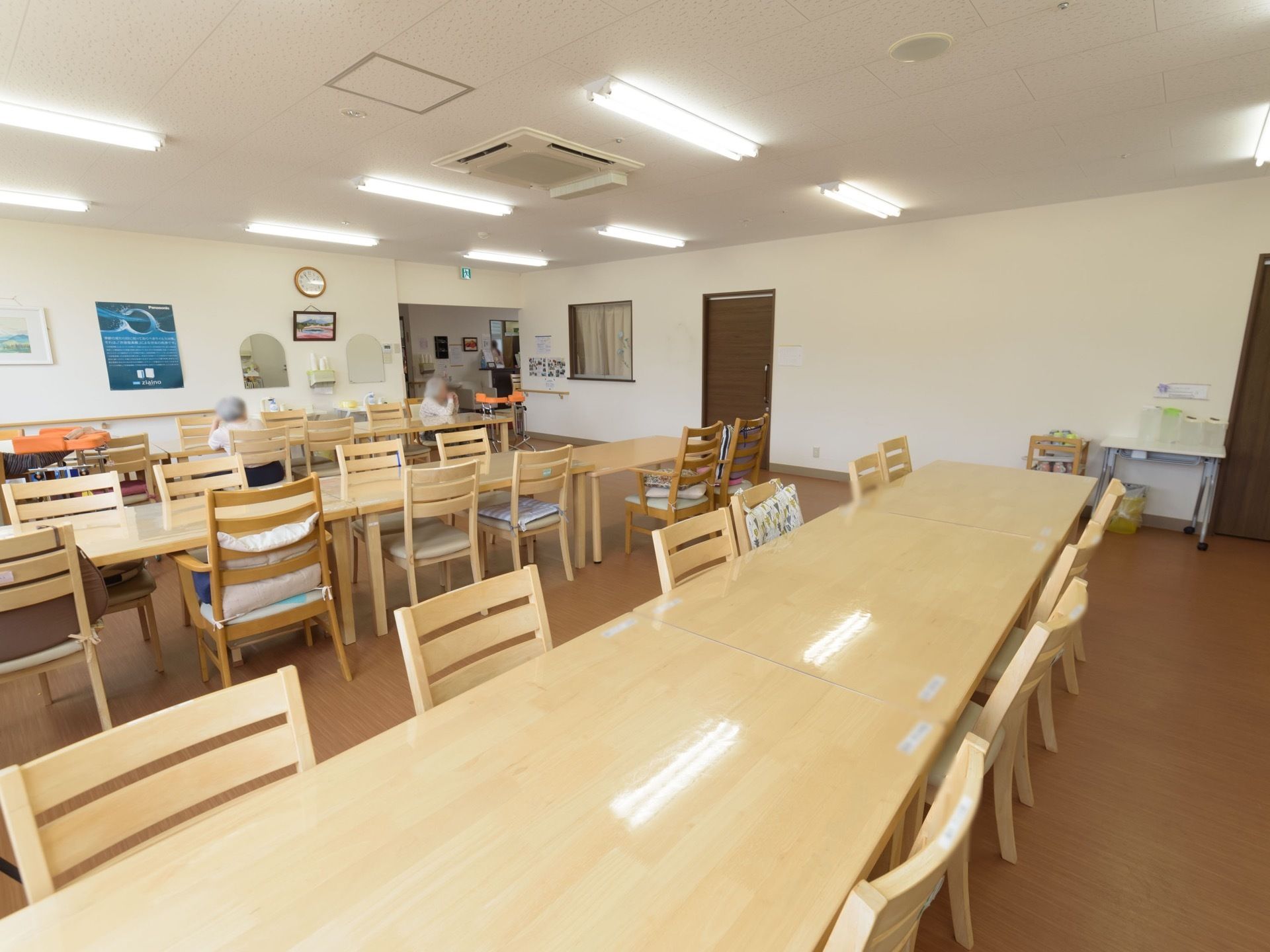 食堂に、たくさんのテーブルや椅子が並んでいる。部屋の一角には洗面台があり、壁には時計やポスターなどが飾られている。