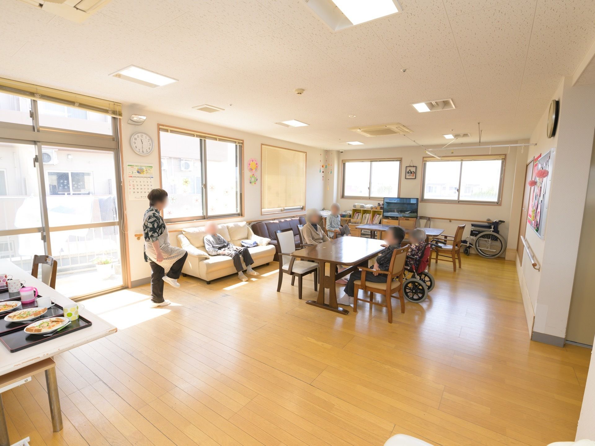 食堂にはガラス扉と複数の窓がある。部屋の中央にはテーブルと椅子が並べられている。壁側にはソファーが置かれている。