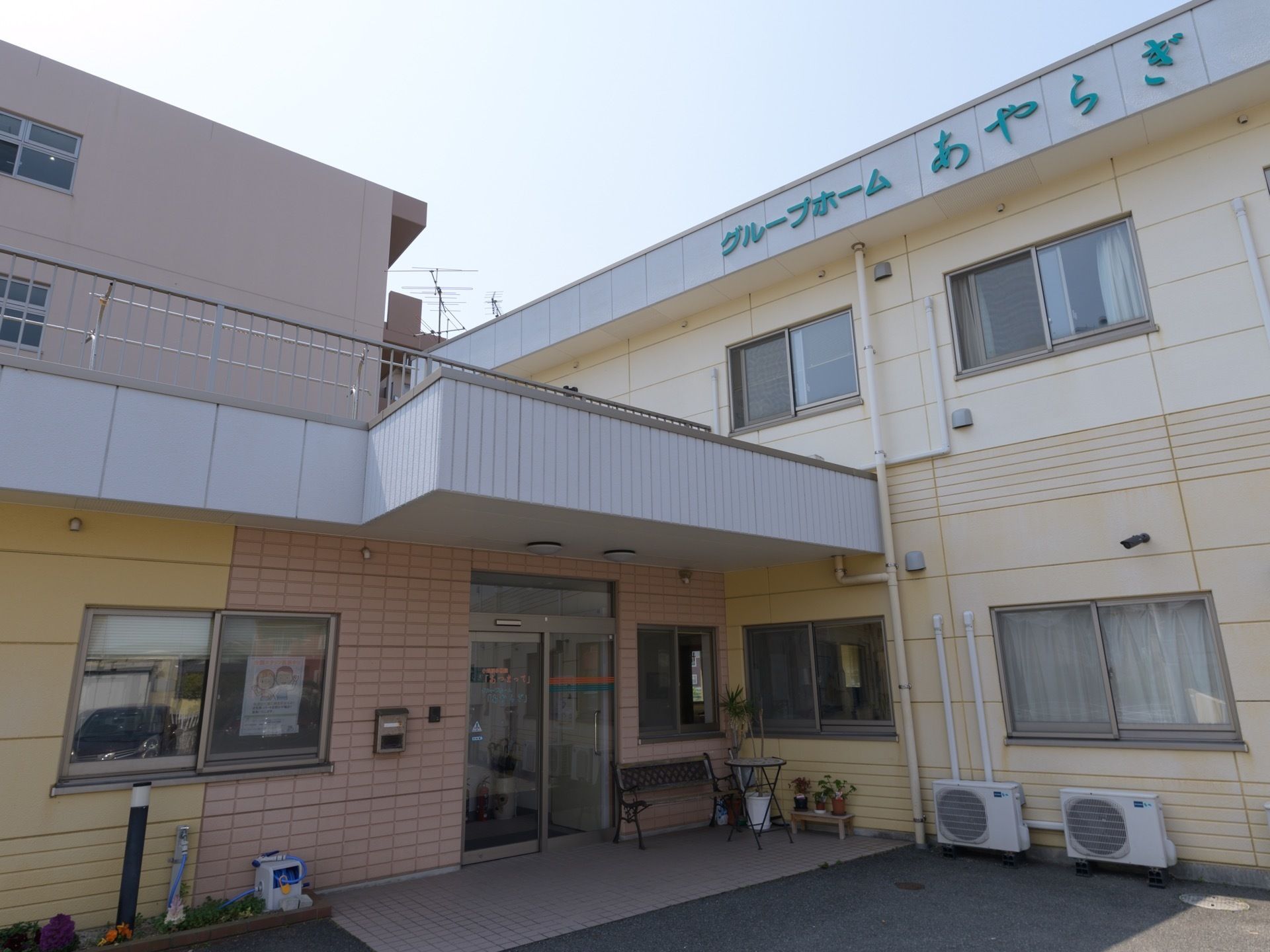 施設は２階建てで、薄い黄色、薄いベージュ色の外観である。玄関の扉はガラス物が設置されており、横にはベンチとテーブルが用意されている。