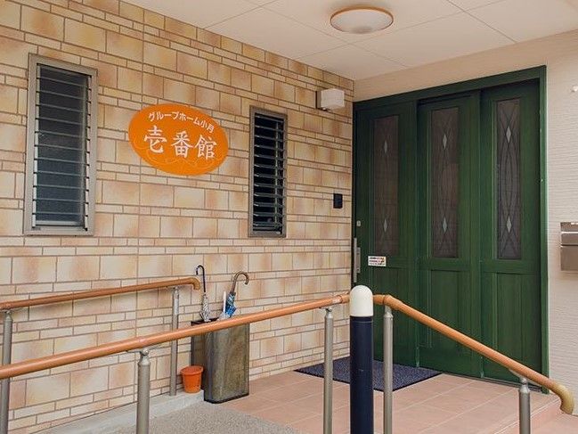 壁に楕円形の看板がある。オレンジ色に白文字で施設名が書かれてある。両わきにルーバー窓が備え付けられている。