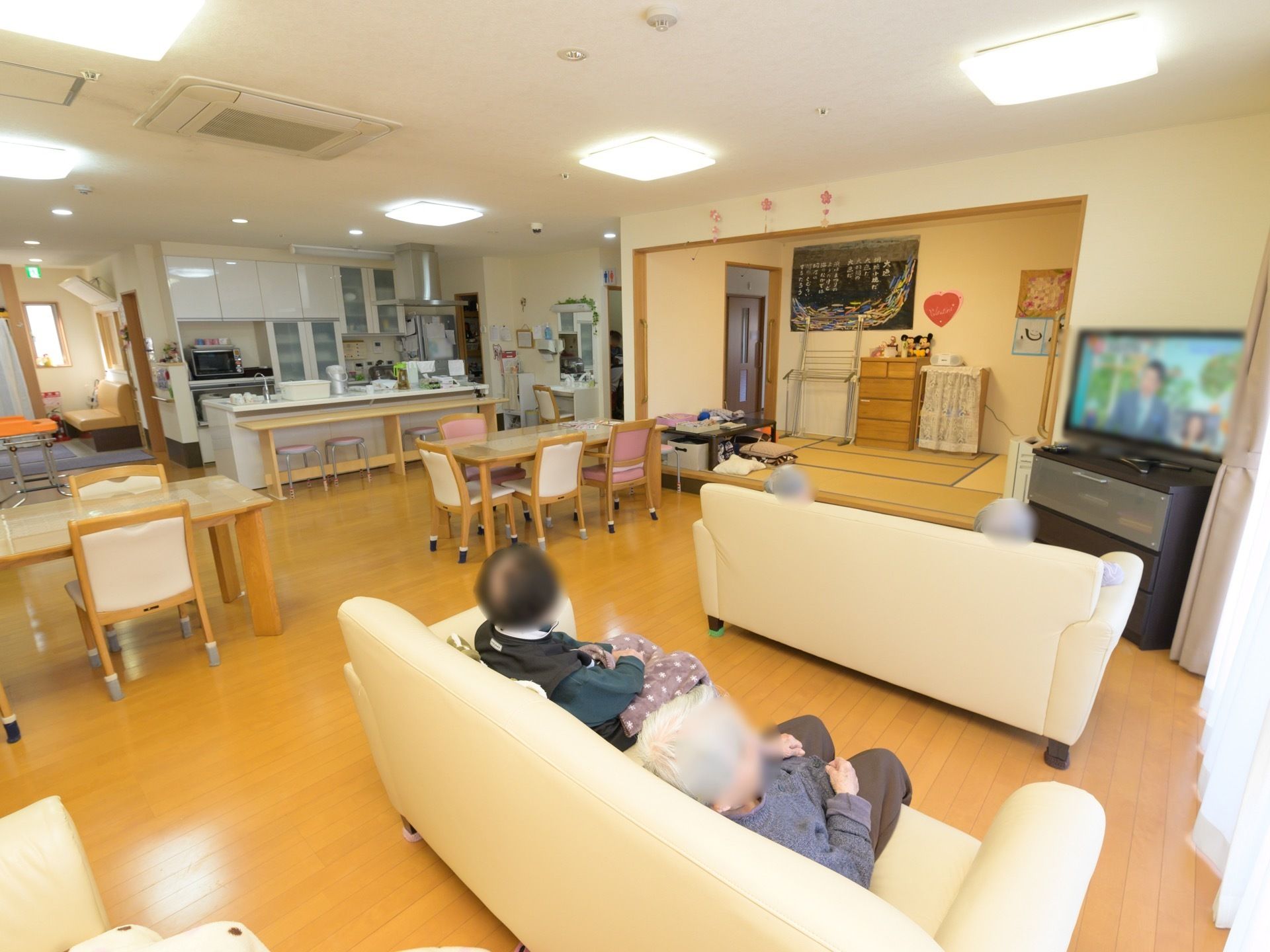 カウンターキッチンや和室スペースが完備された共有スペースである。ソファーに腰掛けテレビをご覧になる入居者様の姿が見える。