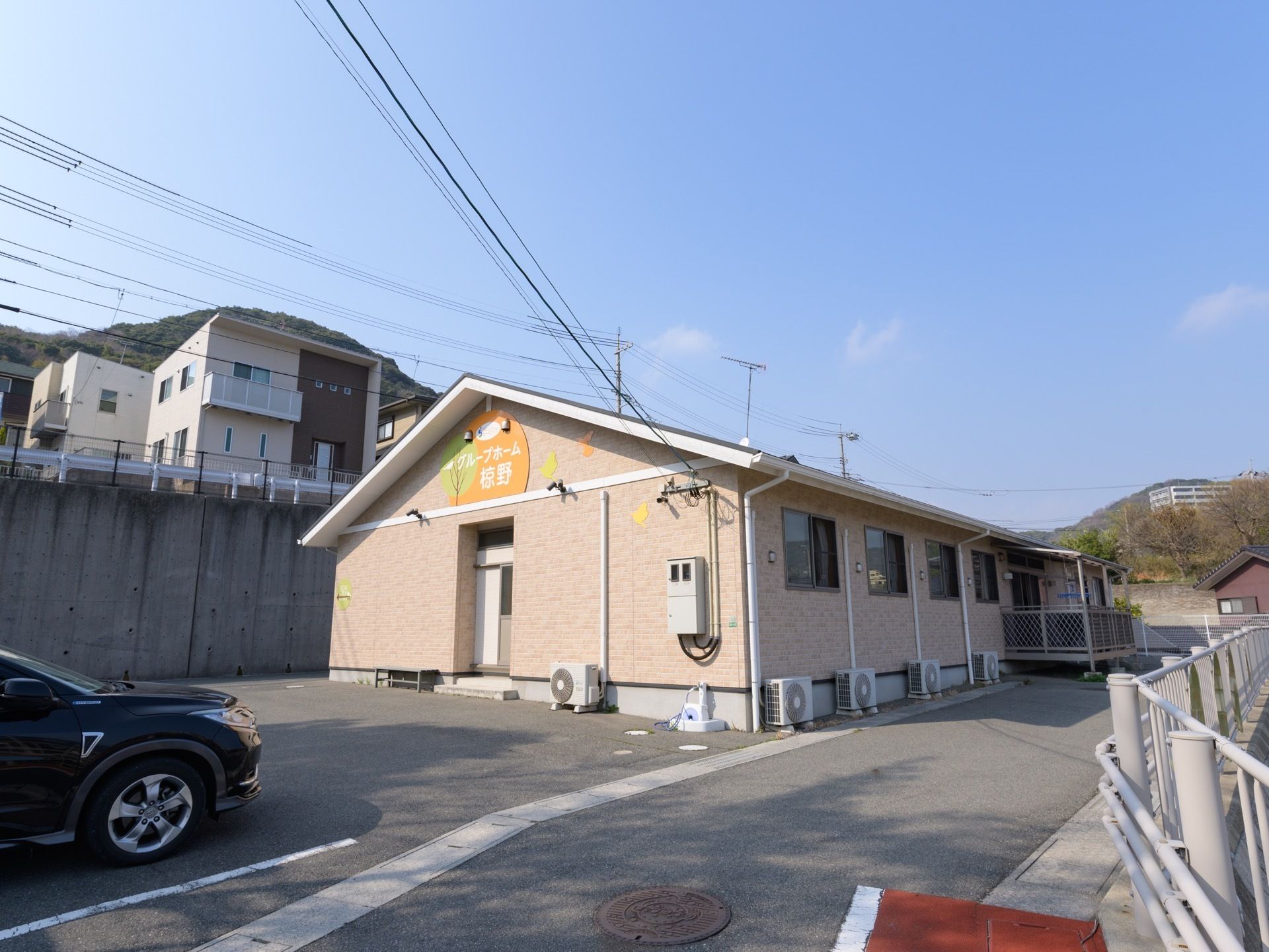 晴れた日に駐車場付近から見た１階建て施設の様子である。完備されたベランダや室外機、駐車場に停車している黒い乗用車が見える。