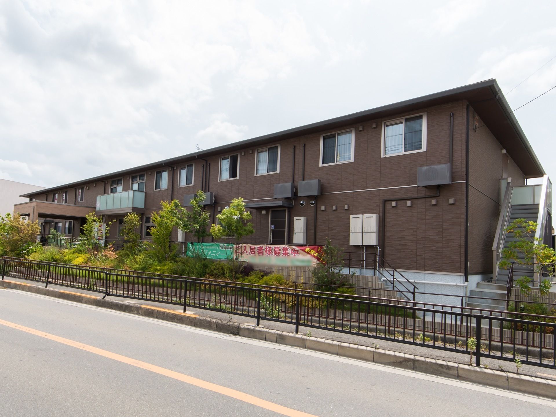 茶色い外壁の2階建ての建物がある。中央あたりに屋根の付いた玄関スペースがあり、端には非常用の外階段も設置されている。