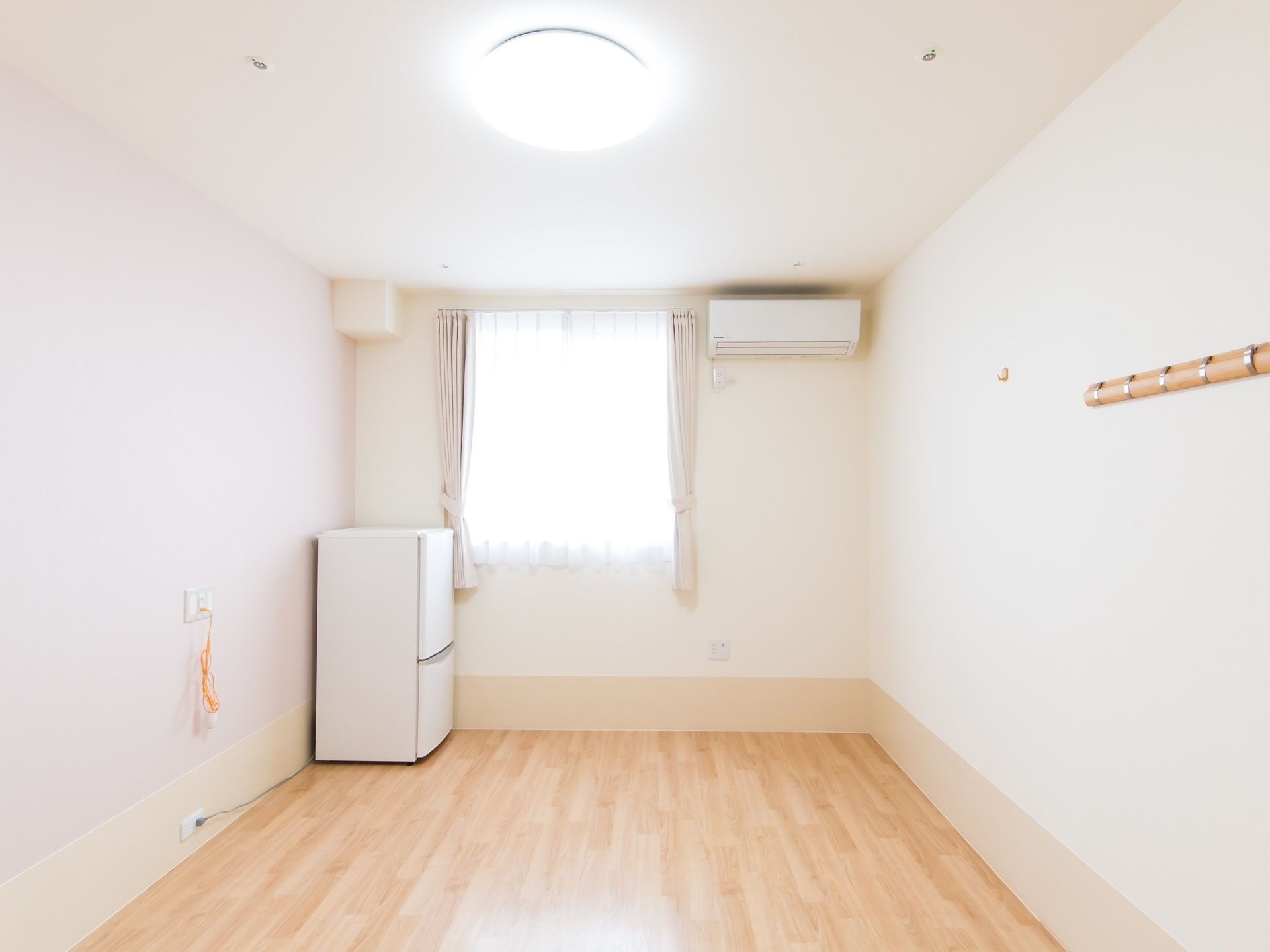 明るいフローリングの居室があり、角に冷蔵庫が設置されている。突き当たりには窓があり、壁にはエアコンが付いている。