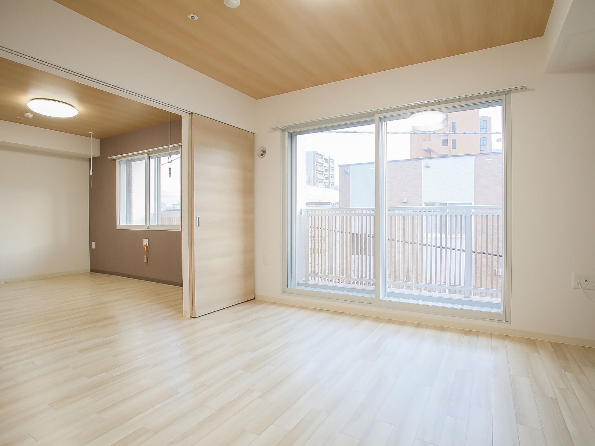 ナチュラルな風合いの居室である。大きめの掃き出し窓の外はベランダで、高めのフェンスがついている。部屋は引き出し式の扉で仕切ることができる。