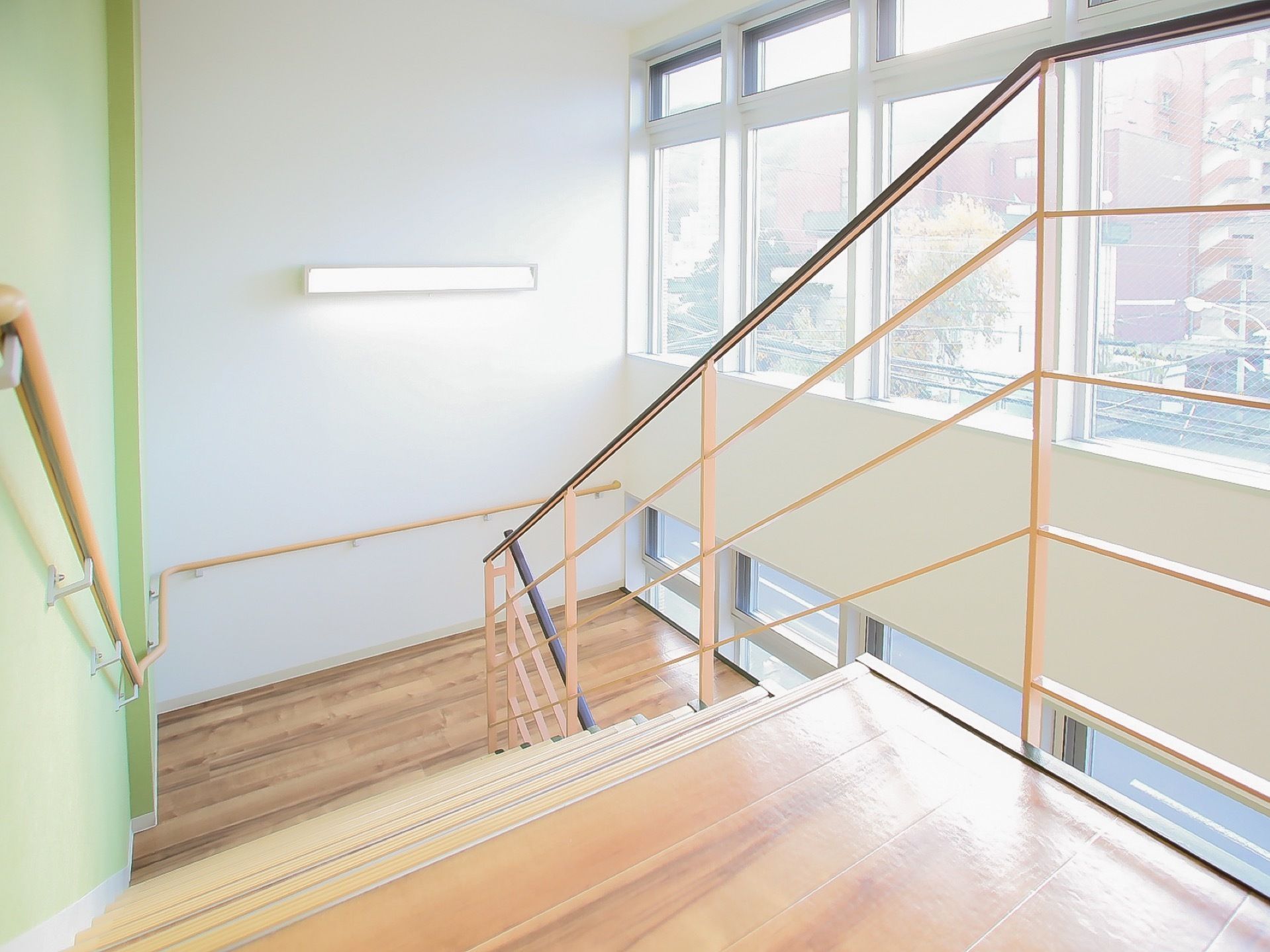 施設建物内にある階段である。壁いっぱいに窓がついていて、光がたくさん差し込んでいる。壁には手すりが設置されている。