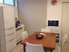 白い壁の室内には、冷蔵庫やミニキッチン、電子レンジなどの設備がある。室内中央に、4人掛けのテーブルが置かれている。