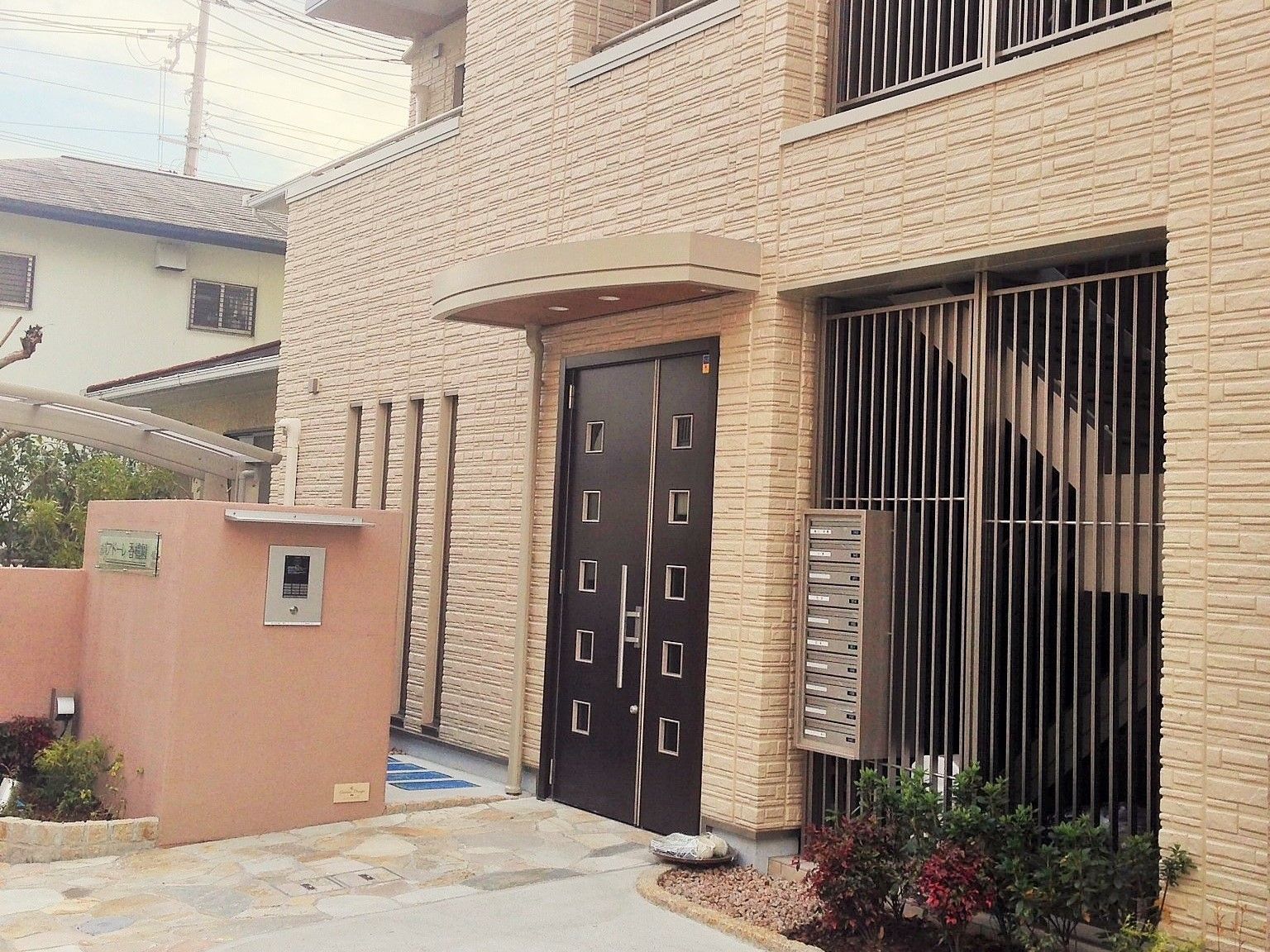 アプローチには石畳が敷かれ、門柱正面には施設名が表示されている。大きめな取っ手が付いた玄関ドアの横には、植え込みがある。