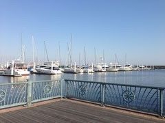 デザイン性のある柵が設置されている、日当たりの良いウッドデッキである。広大な海の上には白いヨットがたくさん並んでいる。