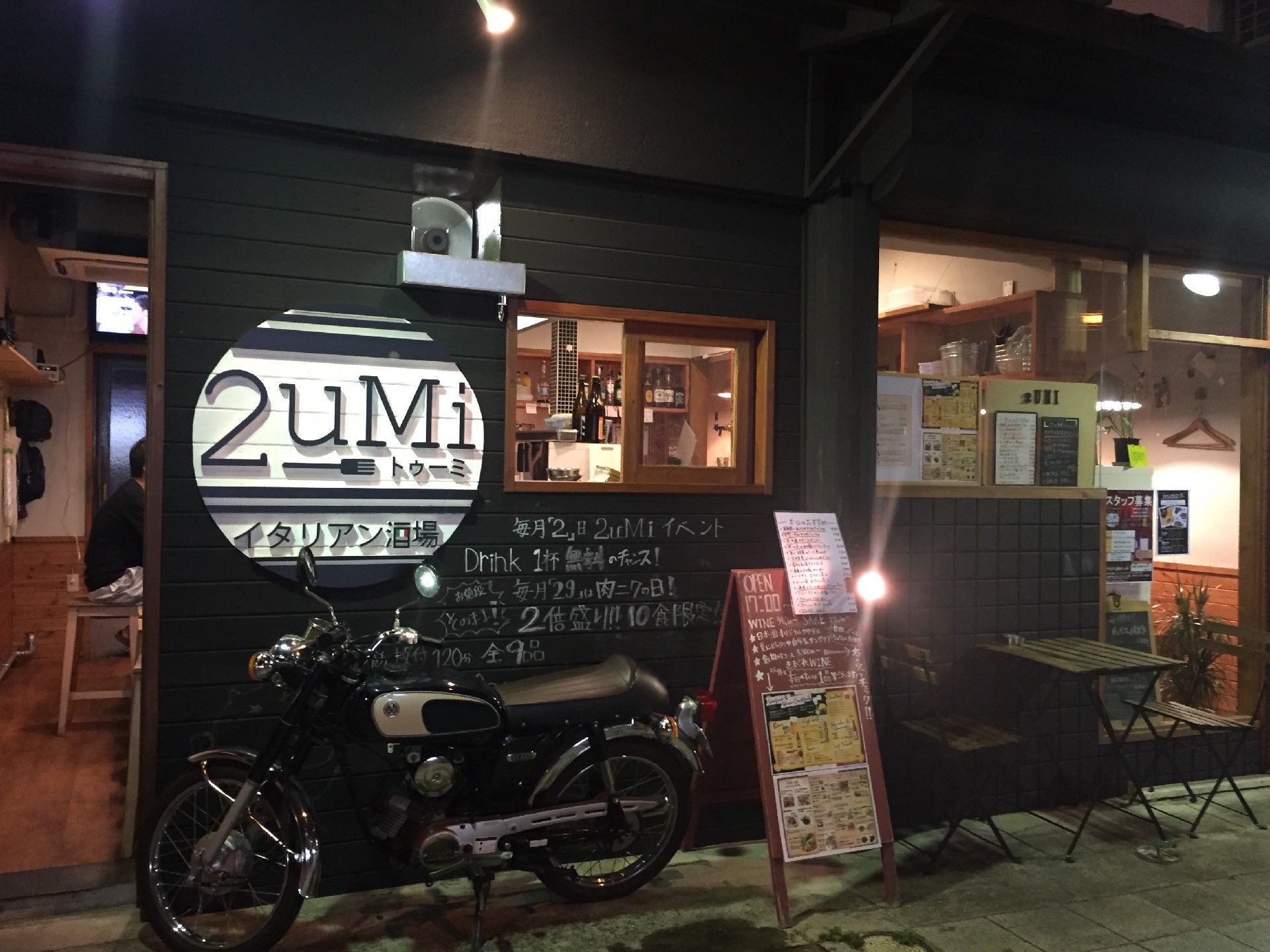 黒い外壁のスタイリッシュなイタリアン酒場である。建物前には、バイクやカフェテーブル、メニューボードが置かれている。