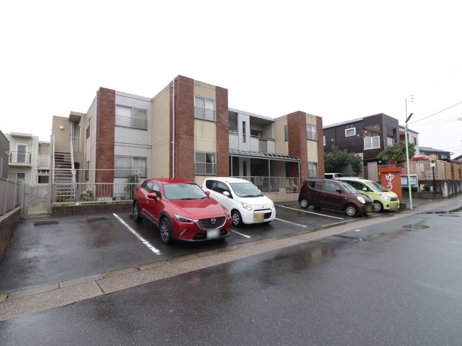 施設の敷地内、道路側が駐車場となっている。フェンスがあり、奥に建物が建っている。左側に非常階段が設置されている。