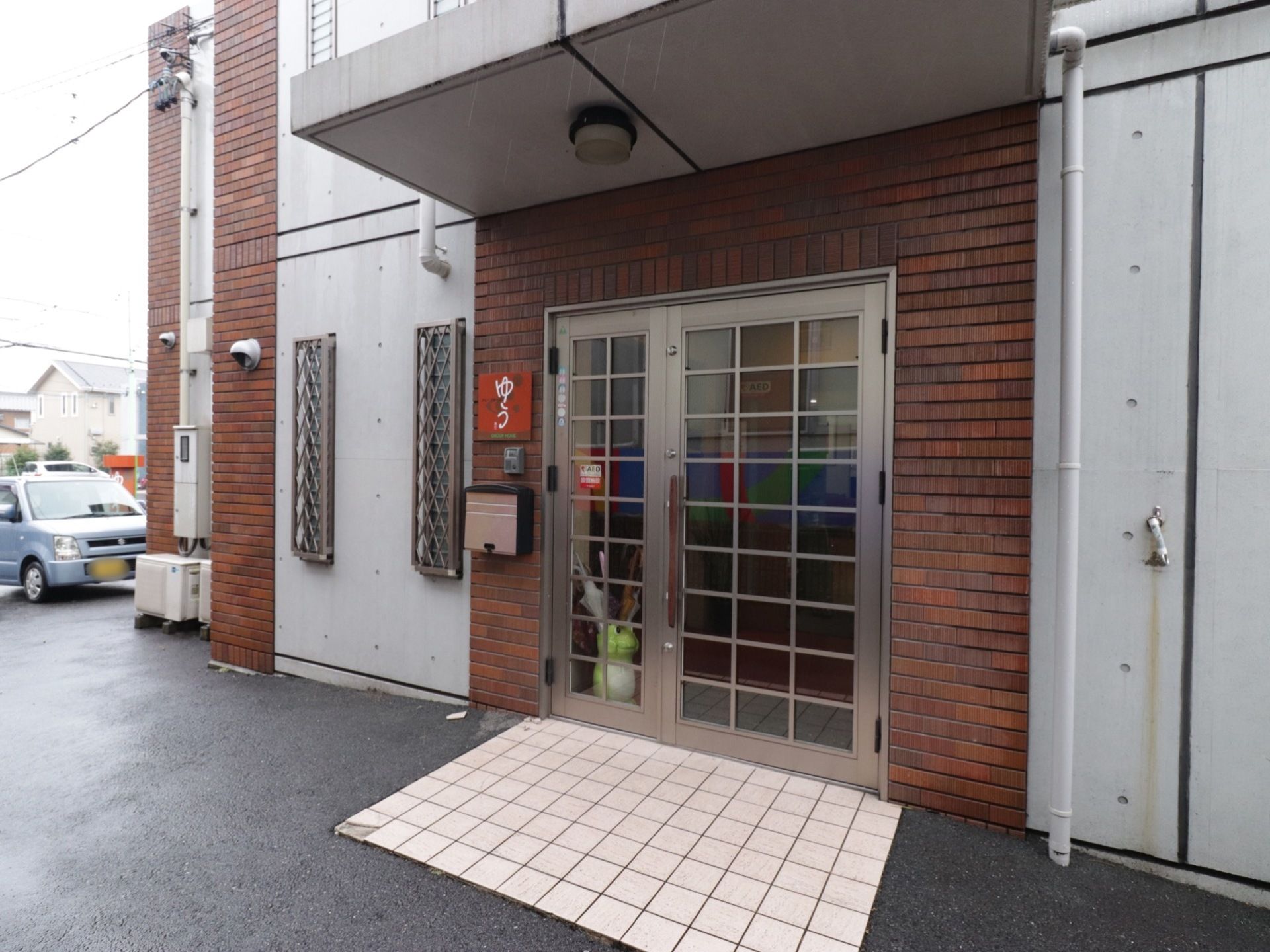 入口は、格子があるガラスドアである。ドア前には、屋根があるので雨の日の出入りに役に立つ。施設内には、AEDを設置していることを表示している。