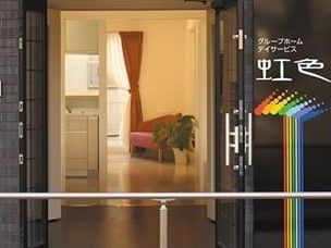 手すりが設置された玄関の扉が開かれ、中の様子が見える。システムキッチンの一部が見え、赤いソファーの横に観葉植物が置いてある。