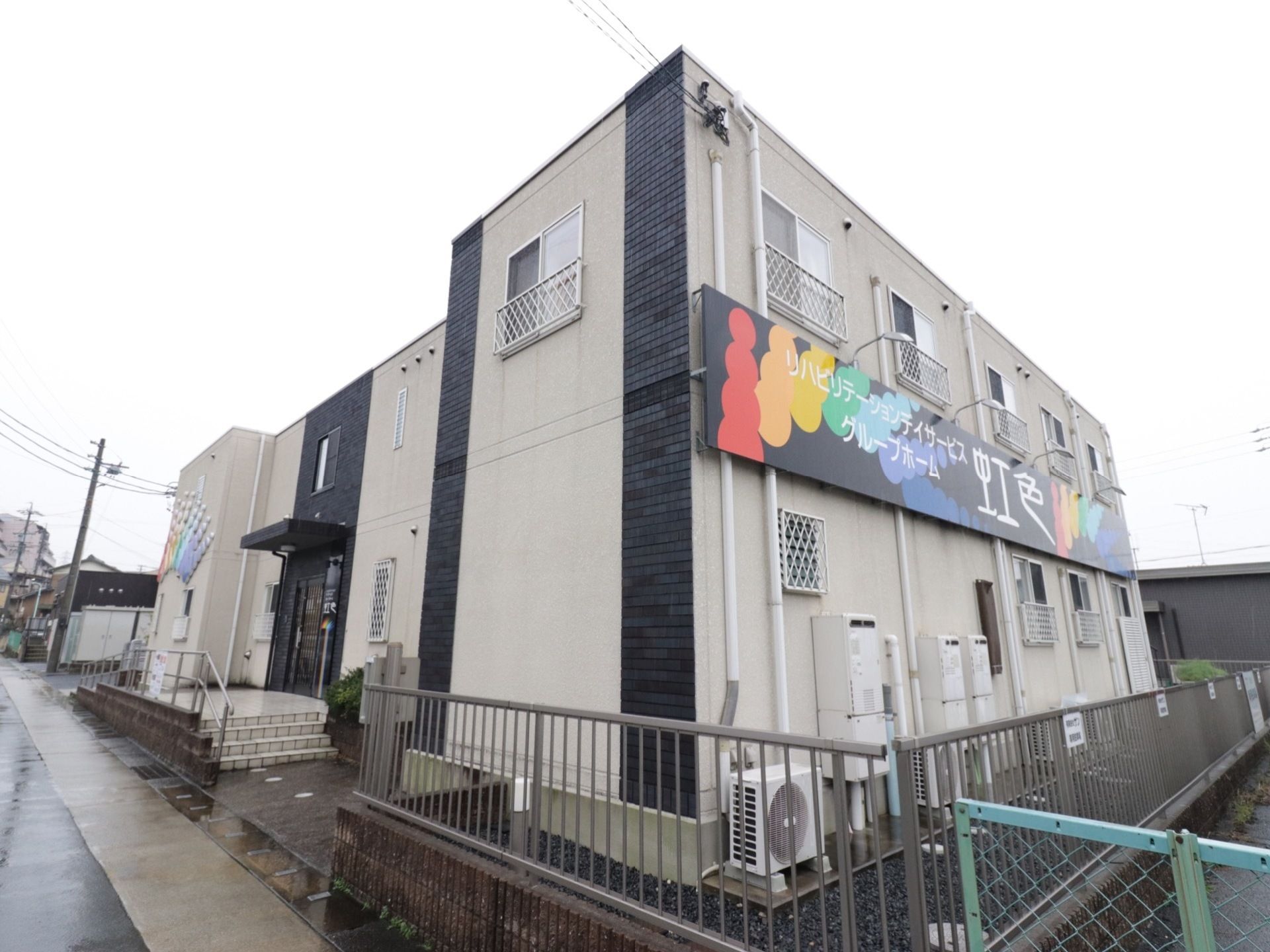 施設の建物は二階建てで、窓には落下防止用のフェンスを設置している。右側の壁面に大きな看板を取り付けている。