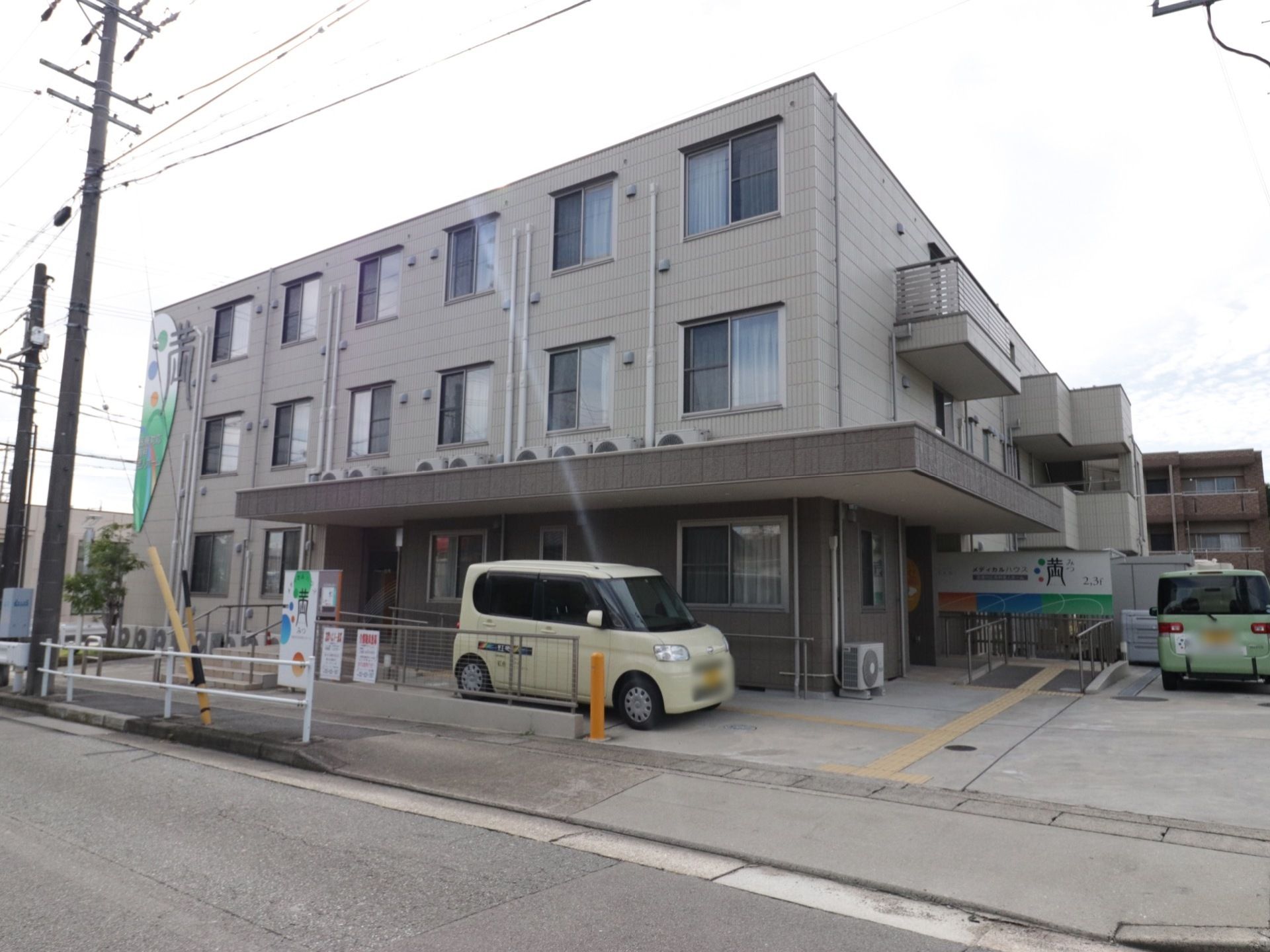 3階建ての建物で、入口全てに屋根と緩やかなスロープが設けられている。施設の送迎車が2台置かれている。