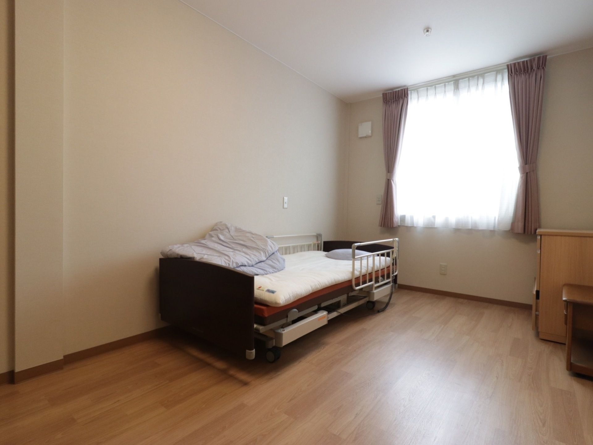 窓にはカーテンがあり、電動介護ベッドと家具が備え付けられている部屋である。ベッドにはお布団も用意されている。