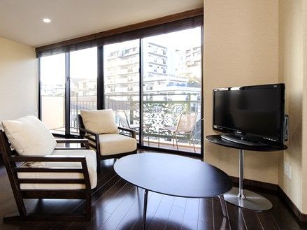 壁際にテレビを備え、前方に丸型のテーブルと１人掛けの椅子を２脚置いている。側面には大きなガラス戸があり、外部にバルコニーを設けている。