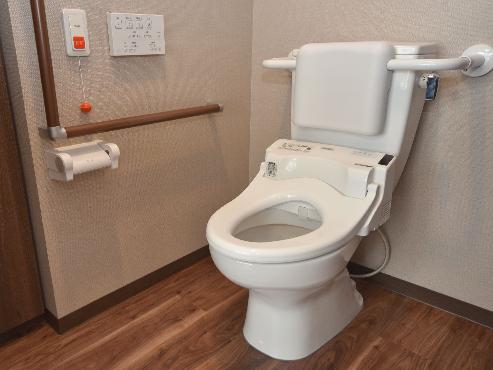 背もたれが取り付けられた水洗トイレは、温水便座が設置されている。左側の壁には呼び出しボタンや温水便座の操作リモコンが用意されている。
