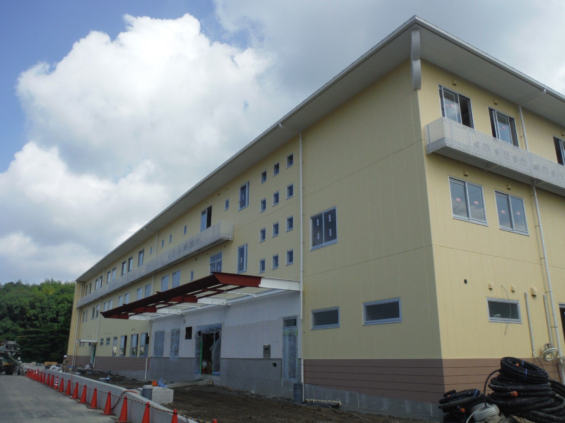 施設の奥には、緑の木々が茂っている。３階建てで横に長い建物で、窓の外にはエアコンの室外機が並んでいる。
