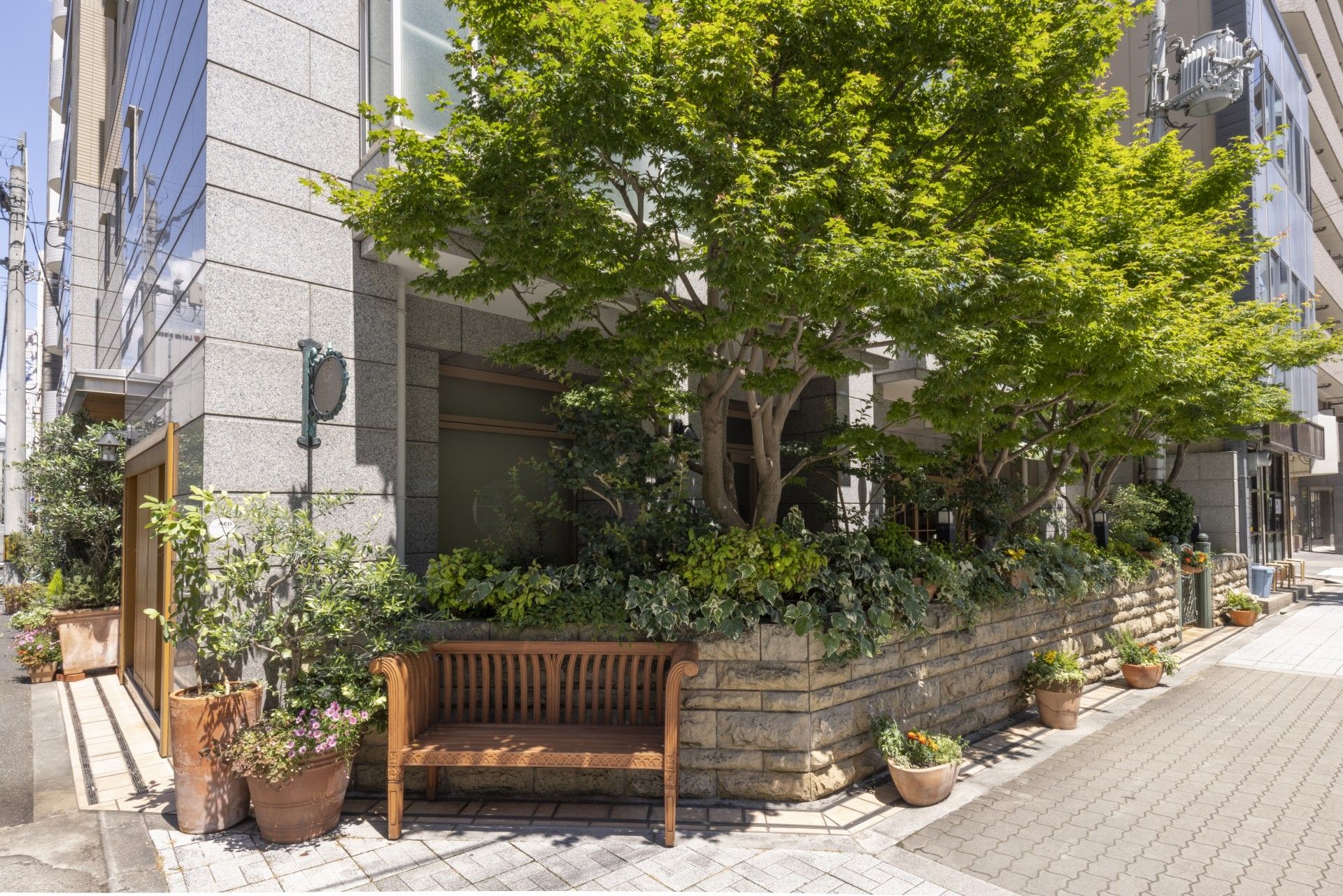 建物の傍に種類の異なる樹木をたくさん植え込み、ブロック塀を設置している。その傍に木製のベンチと花と木の植木鉢をいくつか置いている。