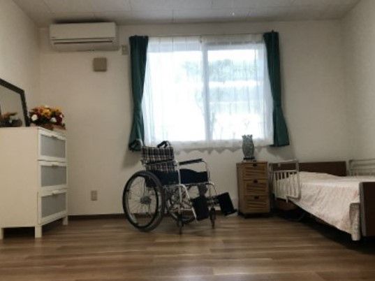 大きな窓のある明るい部屋。落下防止柵がついているベッド、エアコン、２つの収納家具が備え付けてある。車椅子をご利用の方でも利用できる。