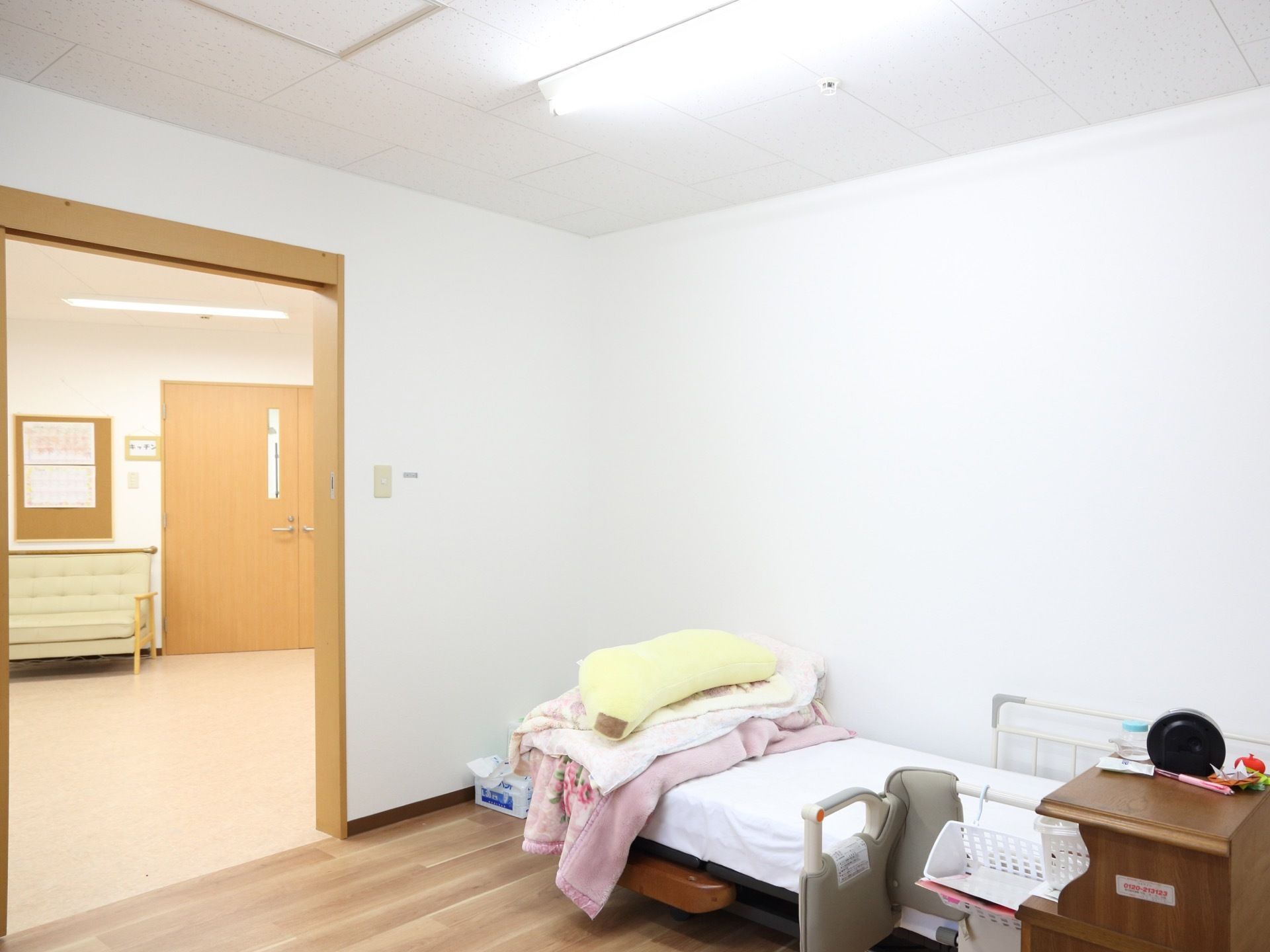 キッチンの扉の向かいには入居者様の居室がある。居室にはベッドがあり、ベッドの上には畳まれた布団が置かれている。