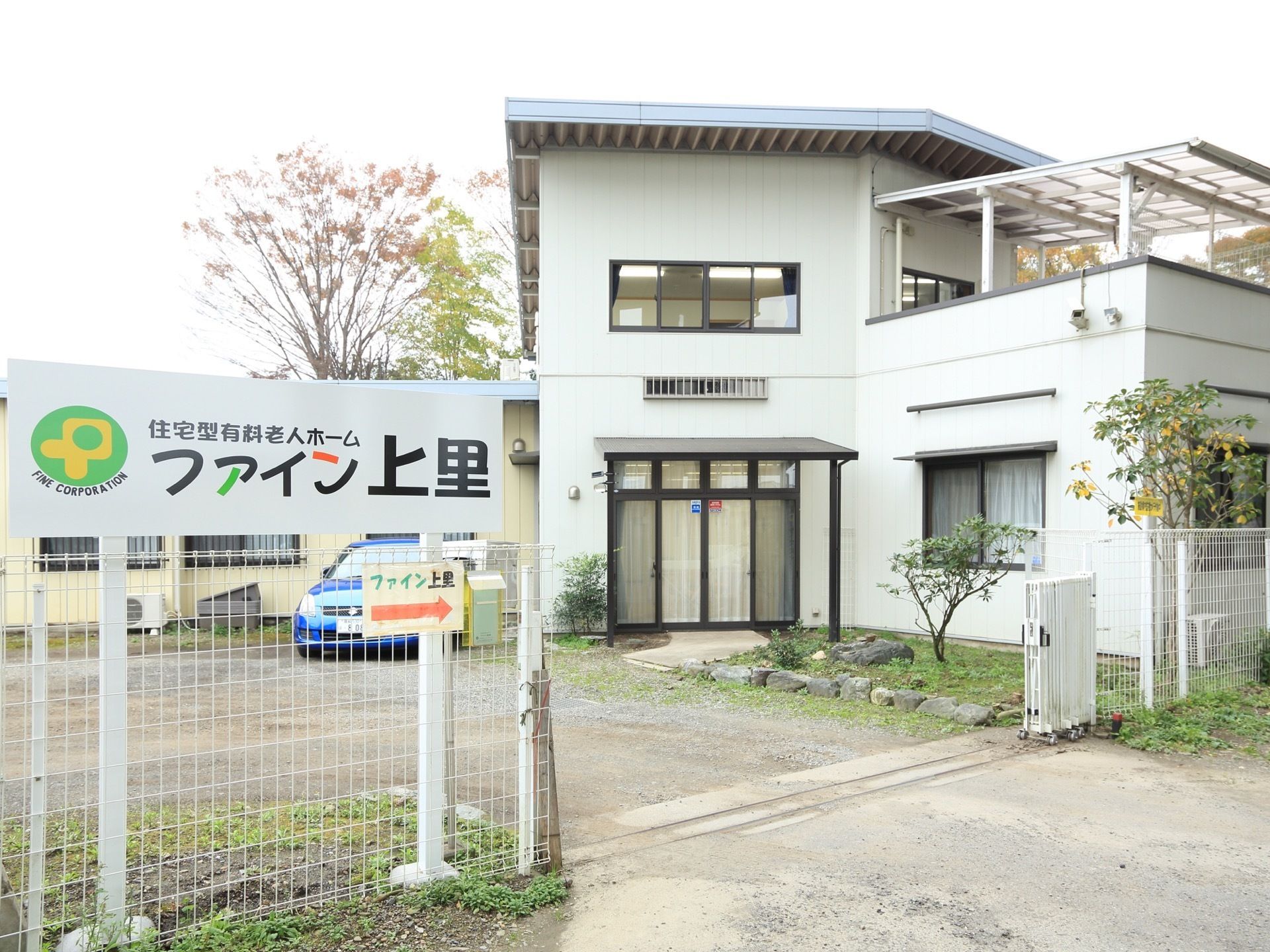 施設は2階建てである。白いフェンスで囲まれた敷地内は全面砂地で、そこには1台のブルーの車が止められている。