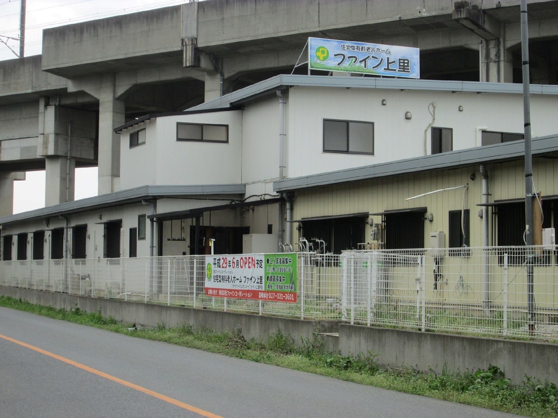 高架下の二階建ての白い建物である。屋上に本施設の看板を立てている。看板は青空の背景に施設の名前を記載している。