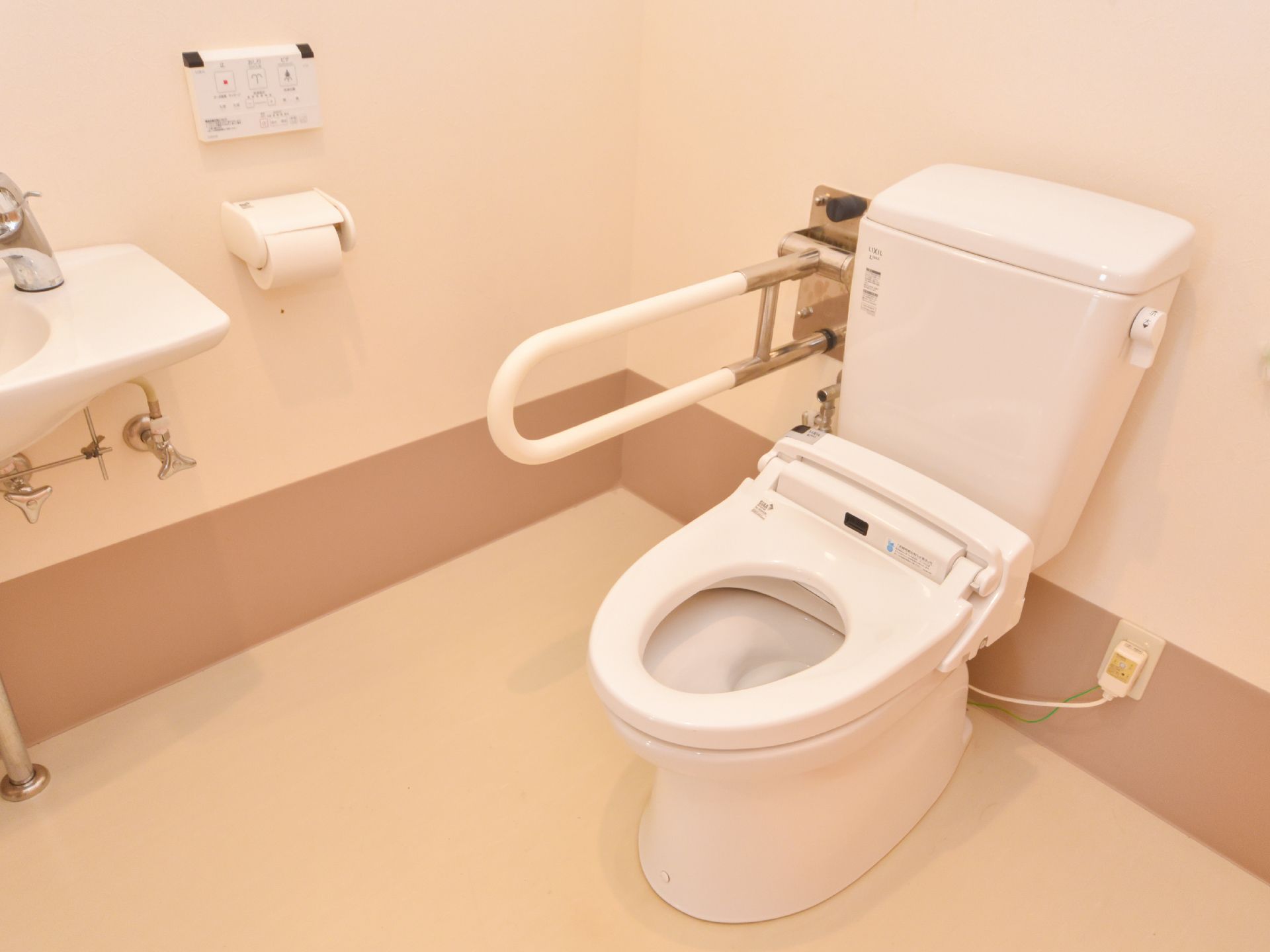 可動式の手すりが設置されている。壁にトイレの操作パネルが取り付けてある。便座のふたのない温水洗浄便座トイレである。