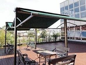建物から繋がる広いウッドデッキのテラスには、緑色の大きな日よけがある。木製のテーブルを囲んでベンチが置いてある。