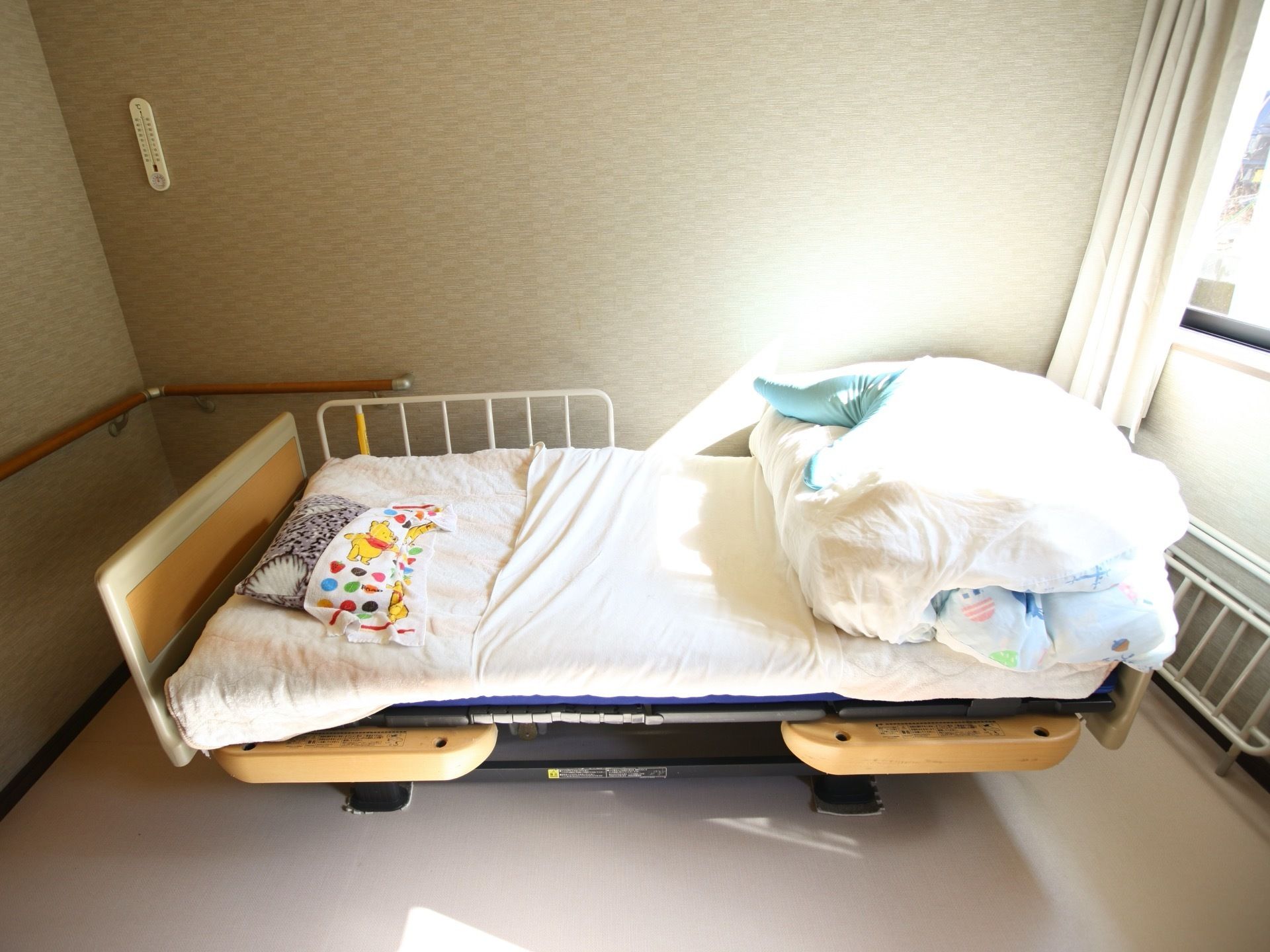 居室のベッドは可動式で、起き上がる際につかまることができるように手すりが付いている。部屋の壁には温度計が掛けられている。