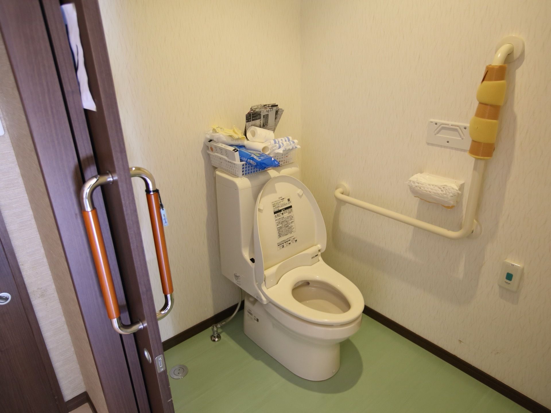 トイレのドアはこげ茶色で、大きな縦長の取っ手がついている。便座の周りの壁にはＬ字型の手すりや緊急用の呼び出しボタンがある。