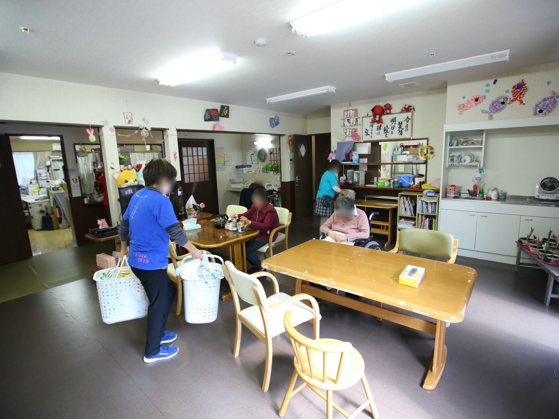 テーブルと椅子が並び、入居者様が座っている。洗濯物が入ったかごを持ったスタッフがテーブルの近くに立っている。