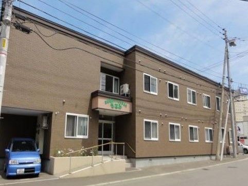 茶色い建物に白い枠の窓が付いている。駐車場から入口へは、スロープが伸びている。玄関の正面は階段である。