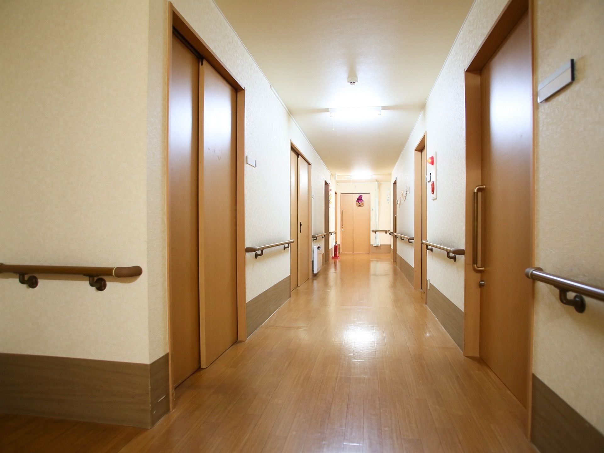 廊下の両側に居室への扉が並んでいる。ドアの横にネームプレートが貼られている。壁に手すりが取りつけられている。