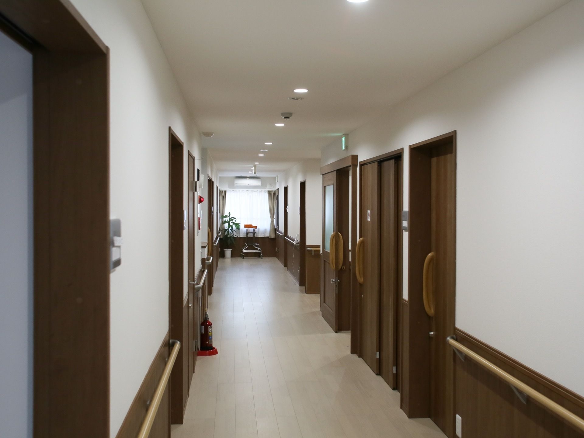通路の床は白いフローリングで、両側には茶色のドアが並んでいる。消火器が置いてあり、天井には火災報知器が設置されている。