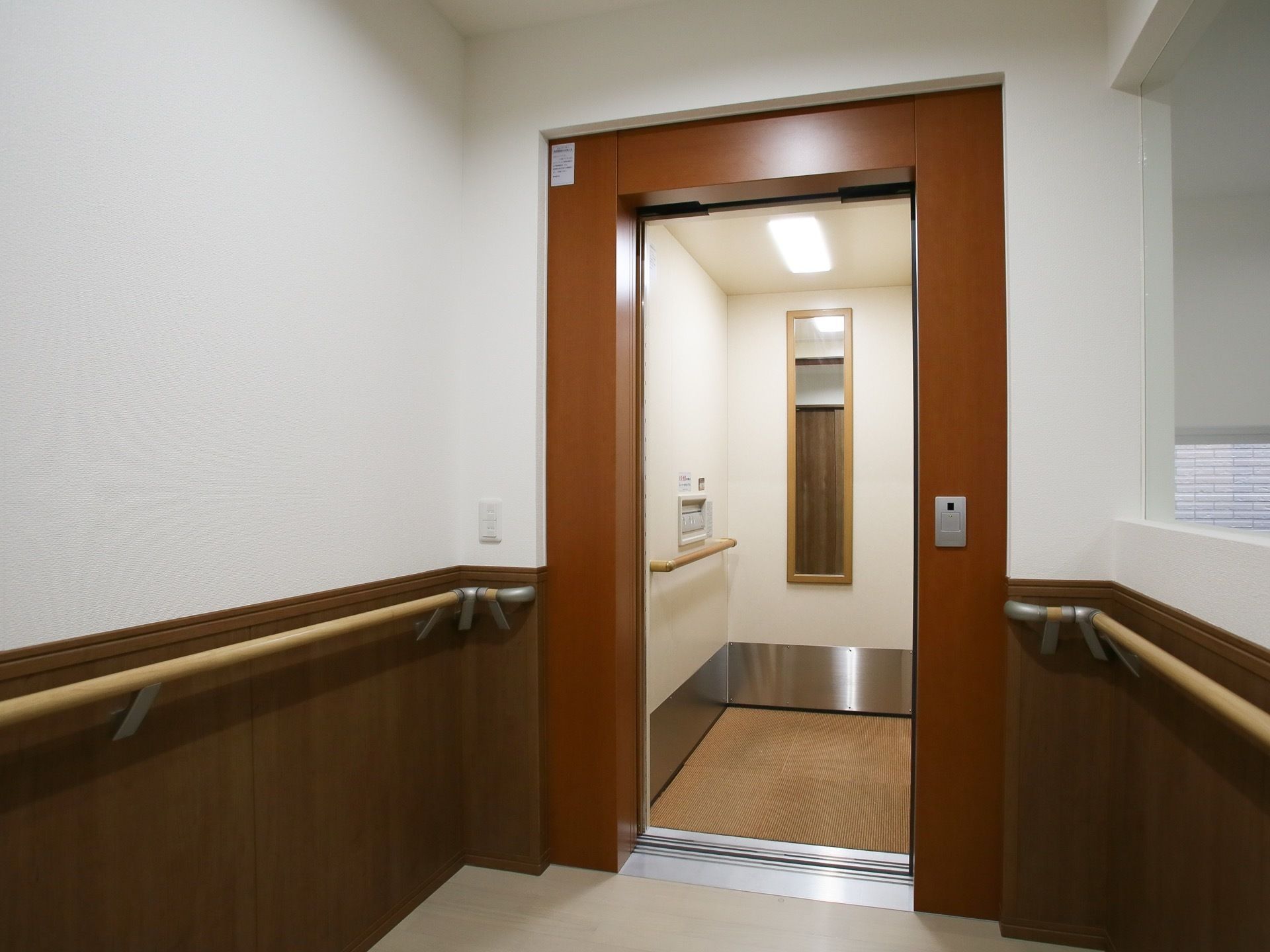 エレベーターの入り口は茶色の木目調になっており、外と中に手すりが備えつけられている。内部は電気がついていて明るく、奥には鏡がついている。