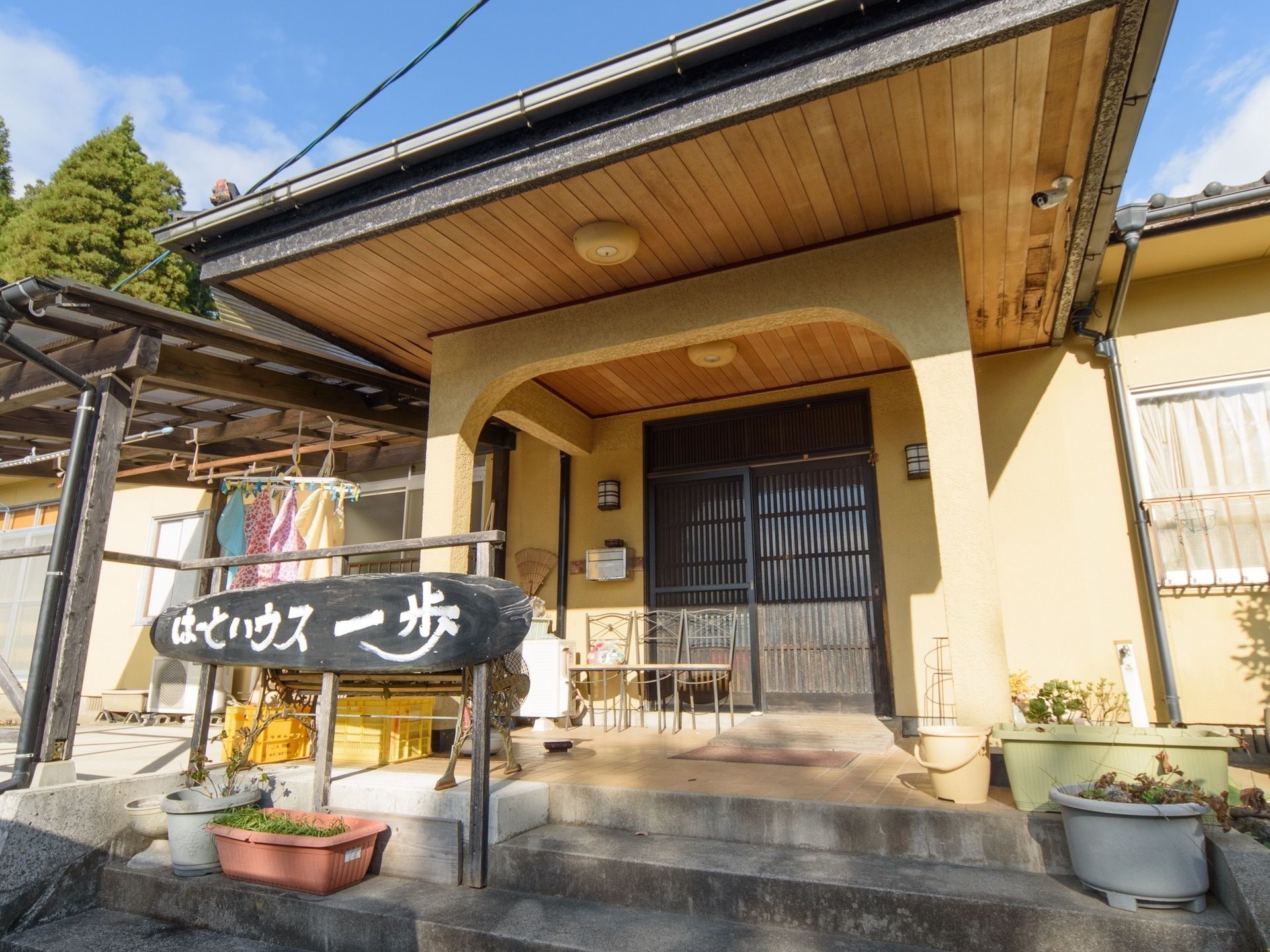 大きなひさしがある玄関。玄関脇には屋根付きのテラスがあり洗濯物は干されている。黒い丸太状の板に施設名を書いた看板が設置されている。