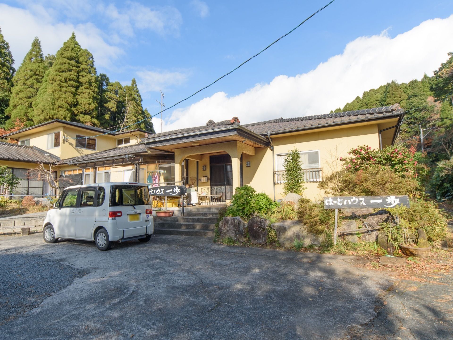 瓦ぶきの屋根にベージュ色の壁の建物がある。手前に駐車場があり、数台が駐車できる。玄関に続く階段がある。