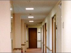 居室が立ち並ぶ廊下である。両サイドの壁には木製の手すりが取り付けられており、通路途中に消火器が置かれている。