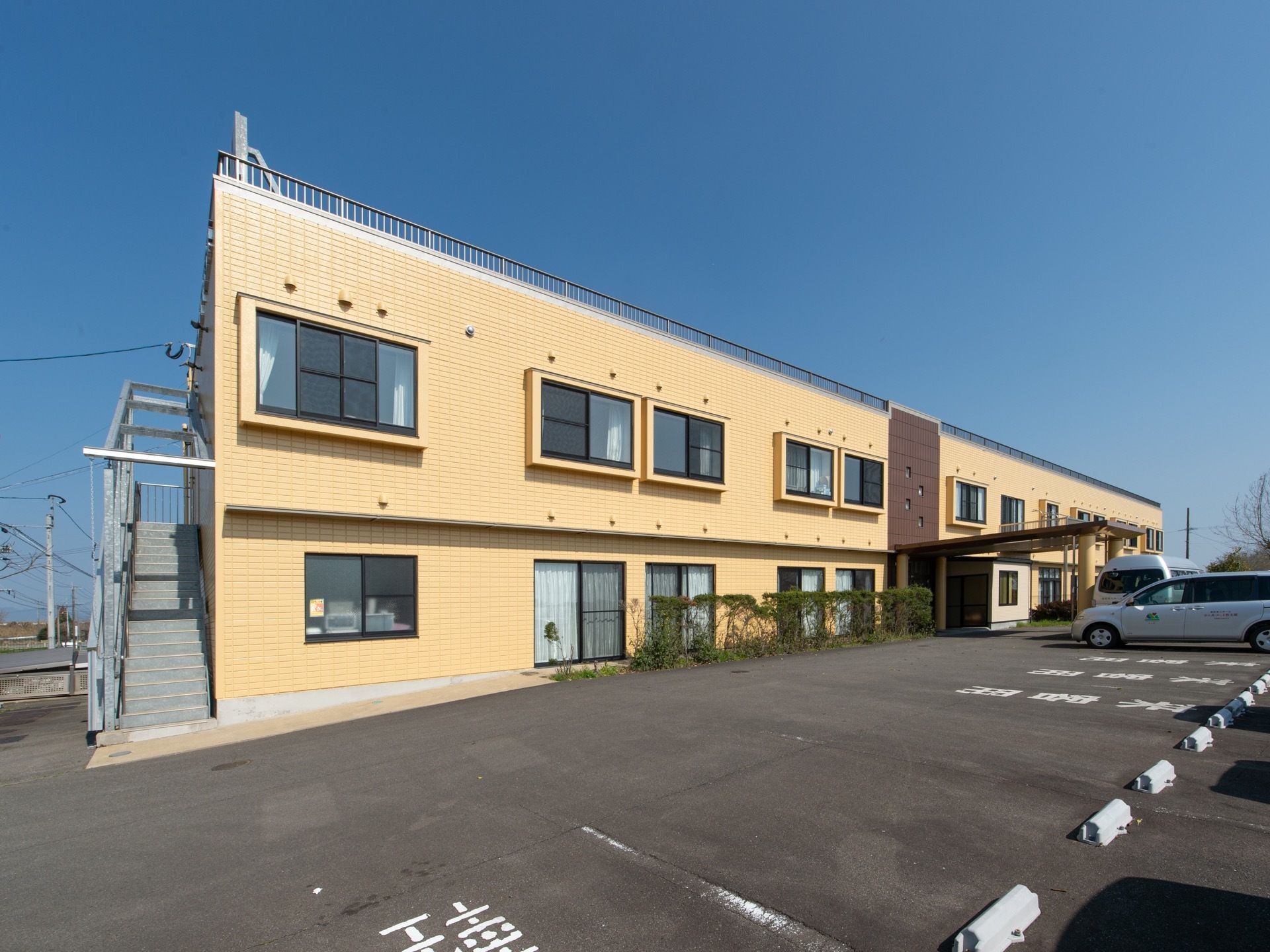 地上２階建ての黄色の外観の施設となっている。敷地内に駐車場があり、軽自動車用や来訪者様専用のスペースが用意されている。